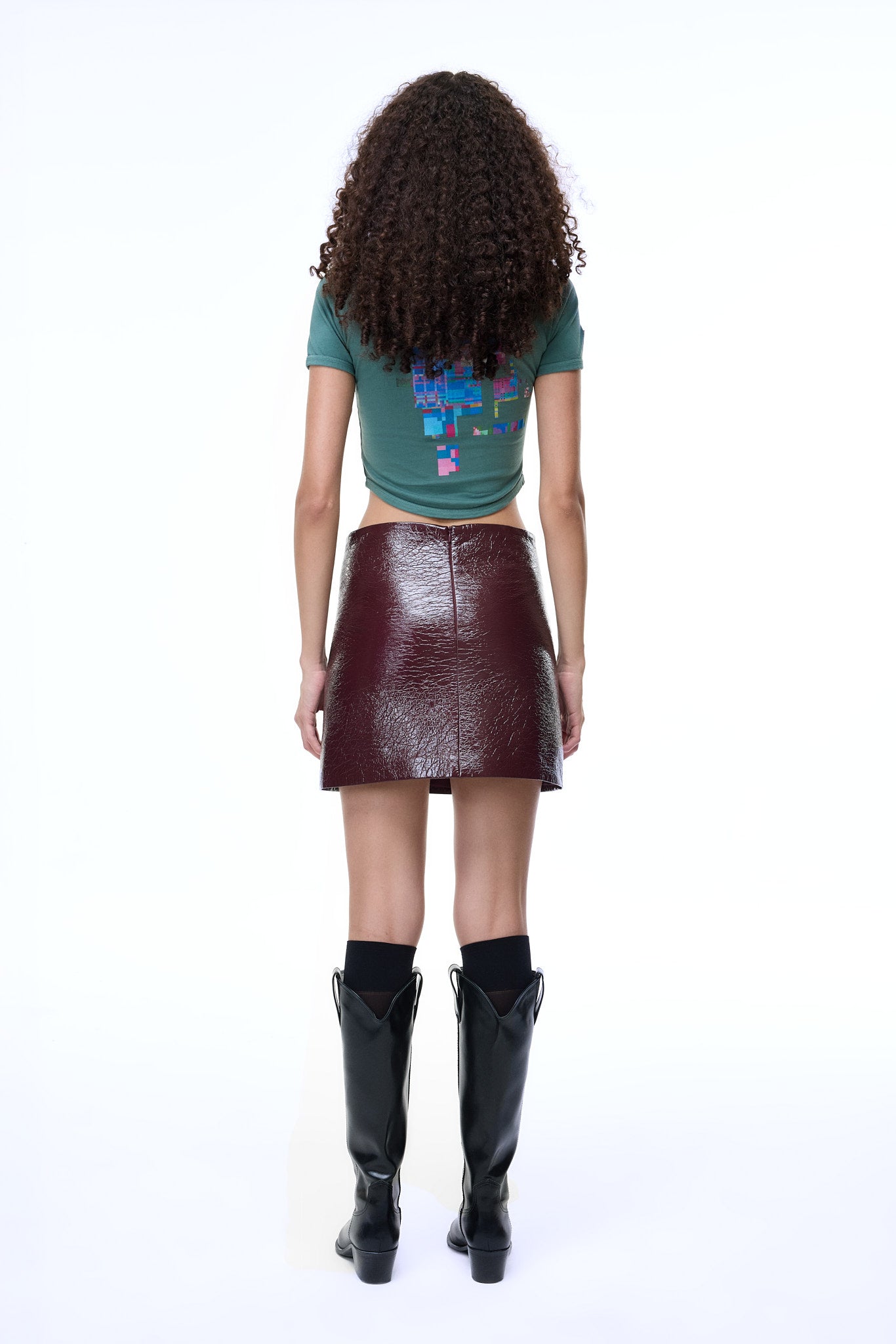Back view of a model with curly hair wearing the GLIX "Lost in Glitch" green crop top baby tee featuring a pixelated colorful back graphic, paired with a maroon patent leather mini skirt and black boots.