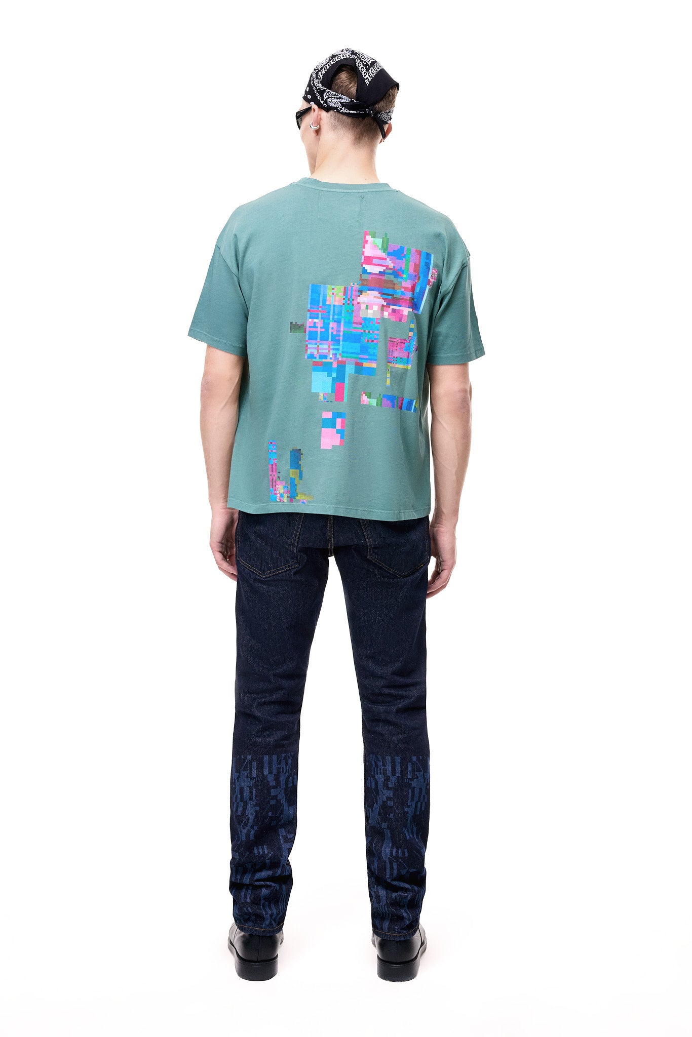 Full body rear view of a male model wearing the green GLIX oversized t-shirt featuring a large colorful pixelated glitch graphic on the back, styled with dark blue patterned jeans and a black and white patterned bandana.