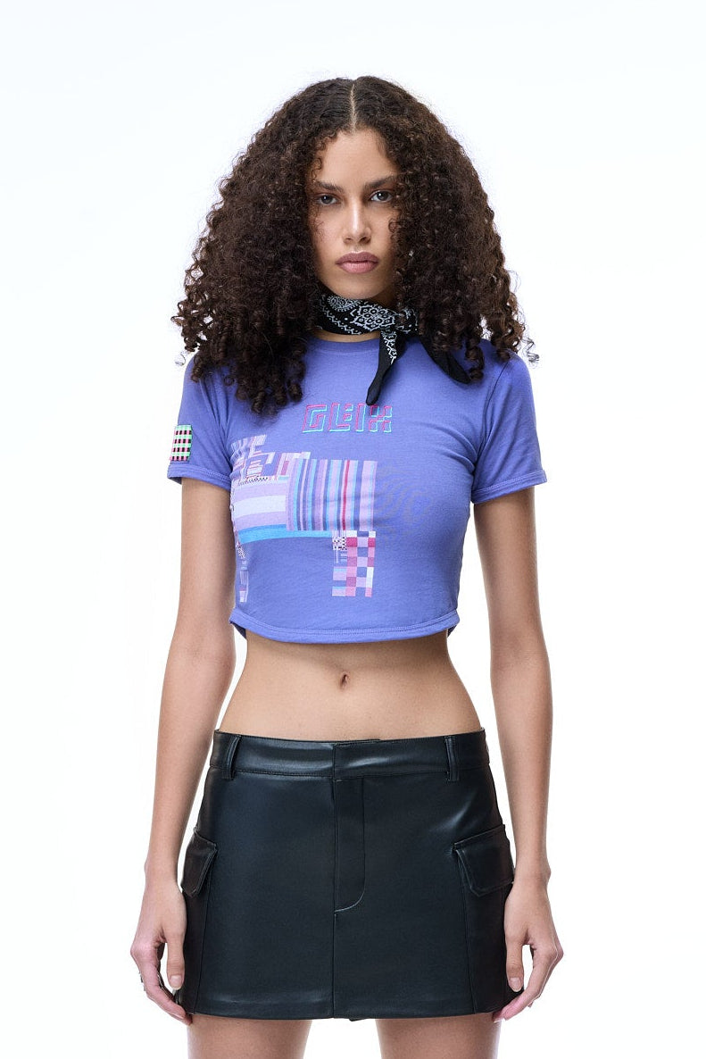 Woman wearing a blue crop top with GLIX text logo embroidery, glitch design print, GLIX tile logo as patch on right arm and black leather skirt on a white background