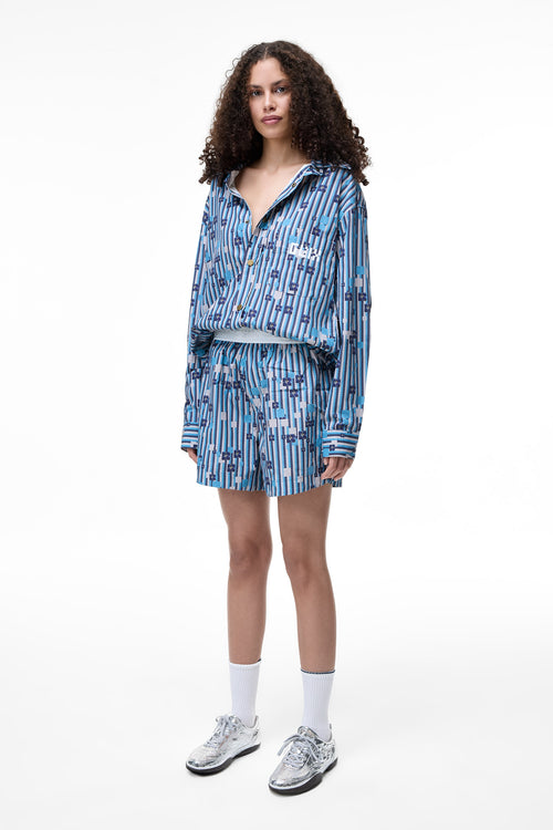 Front lifestyle view of a female model wearing the GLIX "Water Crackle" oversized long-sleeve button-down shirt and matching shorts, featuring a blue and white striped pattern with pixelated floral graphics and a white embroidered logo on the chest pocket.