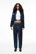 Front view of a female model wearing the GLIX "Totem" dark wash denim crop bomber jacket with a purple and blue glitch-style logo on the chest pocket, styled with matching wide-leg jeans and a white crop top.