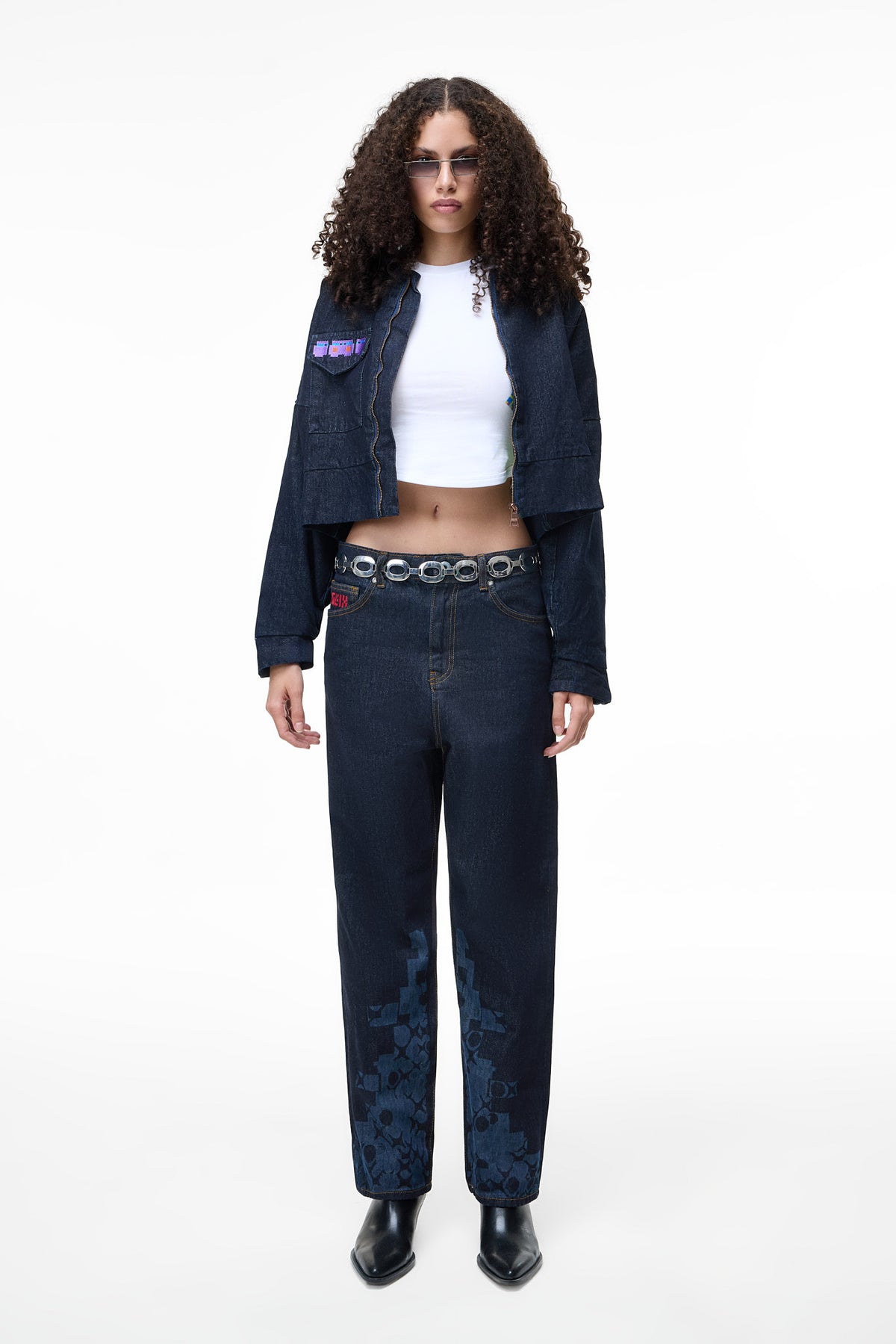 Front view of a female model wearing the GLIX "Totem" dark wash denim crop bomber jacket with a purple and blue glitch-style logo on the chest pocket, styled with matching wide-leg jeans and a white crop top.