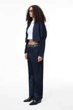 Side profile of a female model wearing the GLIX "Totem" dark wash denim crop bomber jacket, highlighting its cropped length, relaxed fit, and the contrast of the dark denim against the white crop top and matching jeans.