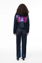 Rear view of a female model wearing the GLIX "Totem" dark wash denim crop bomber jacket, showcasing a large purple, orange, and blue pixelated digital glitch graphic on the back, styled with matching jeans featuring pixelated "V" embroidery on the pockets.