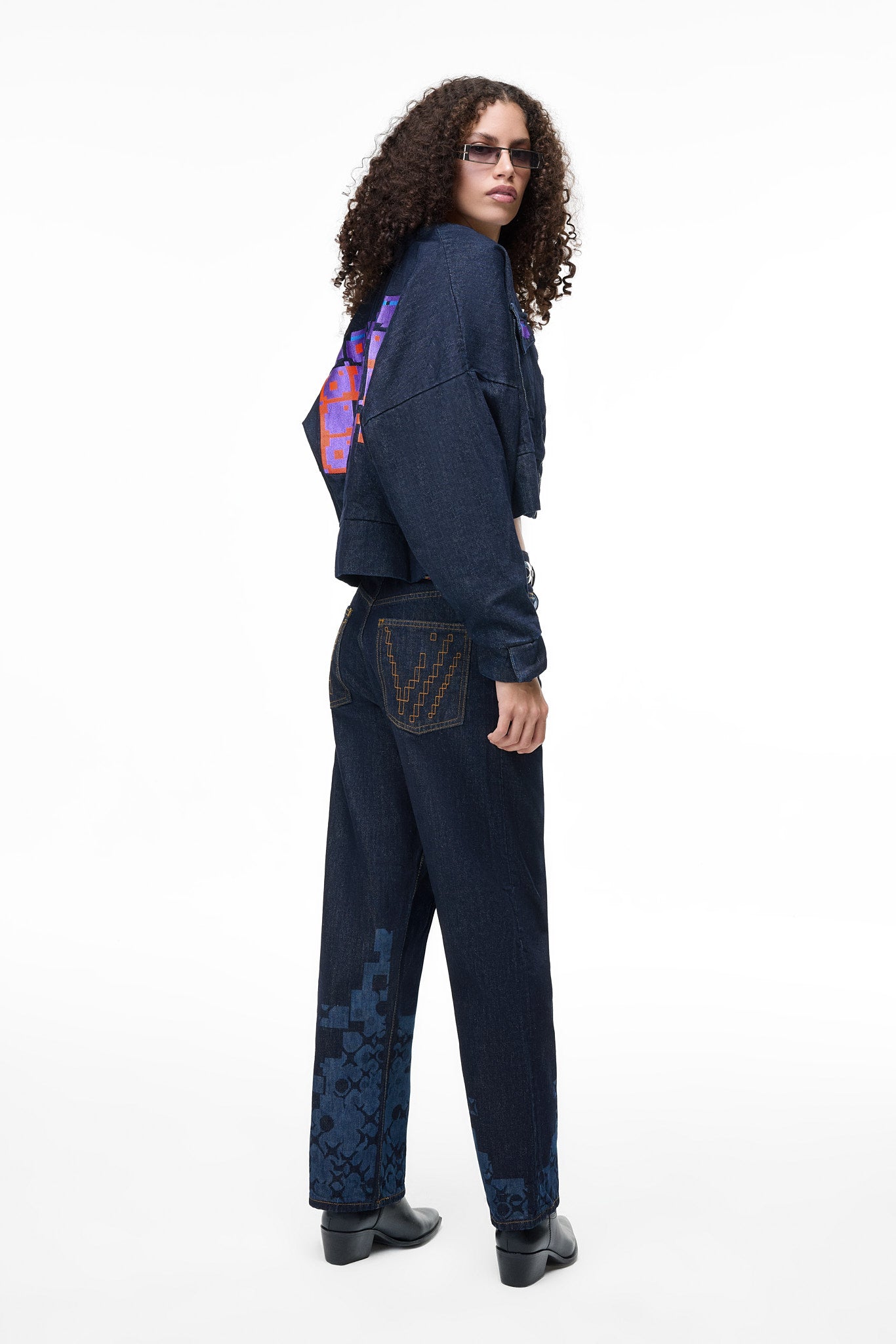 Three-quarter rear lifestyle view of a female model in the GLIX "Totem" dark wash denim crop bomber jacket, showing the side profile of the large purple and orange pixelated back graphic and the "V" embroidery on the matching jeans' back pocket.