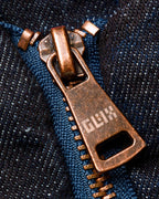 Macro close-up of the GLIX "Totem" denim bomber jacket's copper-toned metal zipper pull, featuring the pixelated "GLIX" logo engraved on the tab, set against dark wash denim fabric and blue zipper tape.