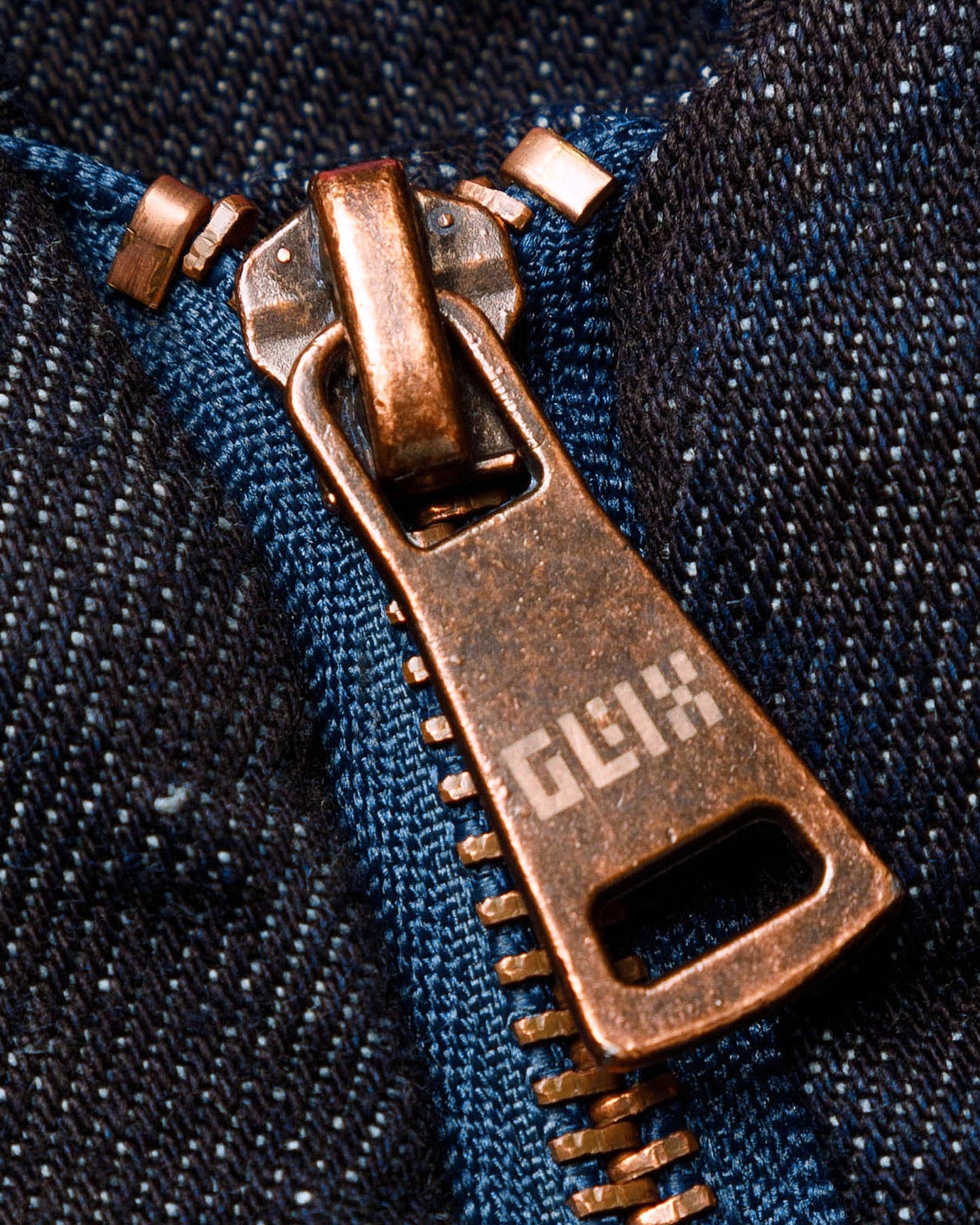 Macro close-up of the GLIX "Totem" denim bomber jacket's copper-toned metal zipper pull, featuring the pixelated "GLIX" logo engraved on the tab, set against dark wash denim fabric and blue zipper tape.