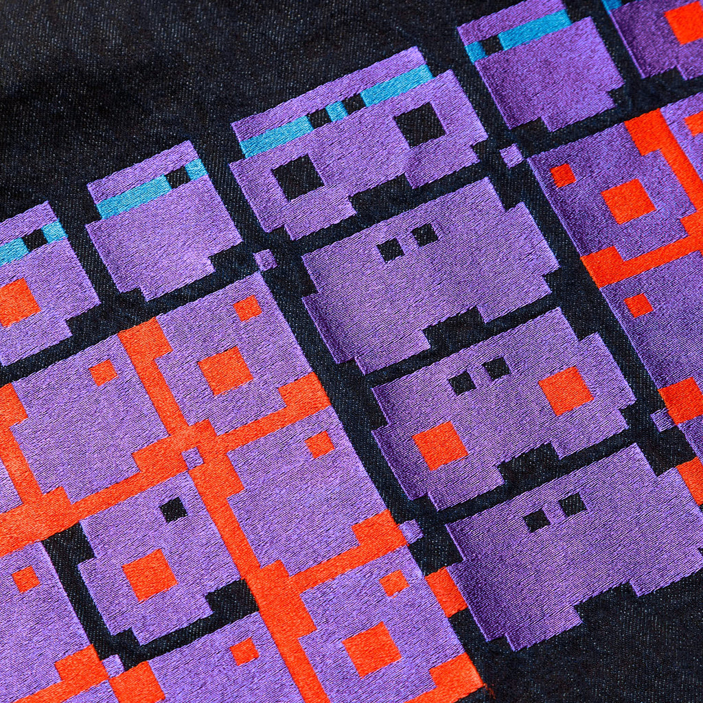 Macro close-up of the GLIX "Totem" chest pocket, featuring three pixelated glitch-style character logos embroidered in purple, light blue, and bright orange thread on dark wash denim.