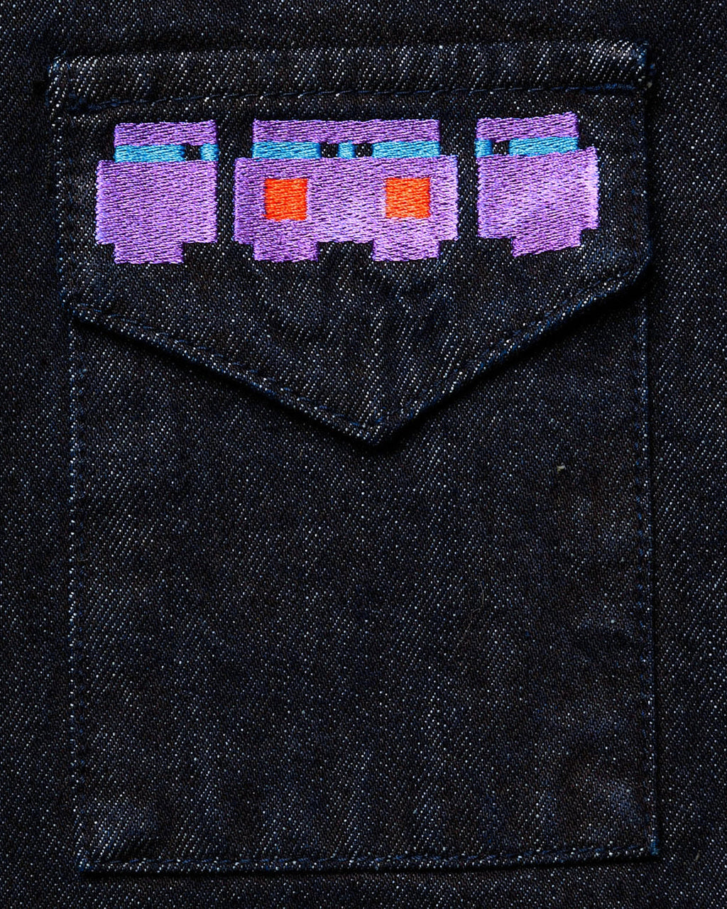 Macro close-up of the large "Totem" back graphic on dark wash denim, featuring high-density pixelated embroidery in vibrant purple, orange, and blue threads.
