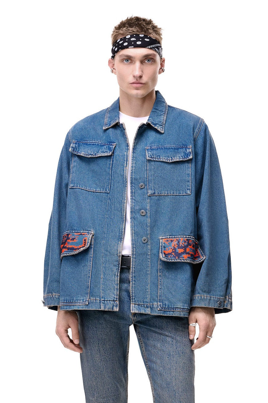 Front upper-body view of a male model wearing the GLIX "Coral Biome" oversized denim jacket, highlighting the two chest pockets and the distinctive orange and blue digital glitch pattern on the lower flap pockets.