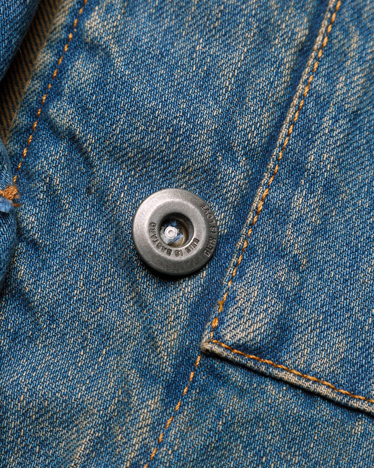 Macro close-up of a silver-toned metal button on the GLIX "Coral Biome" denim jacket, featuring the engraved "GLIX IS LOVE / GLIX IS BASTARD" slogan and orange contrast stitching on blue denim.