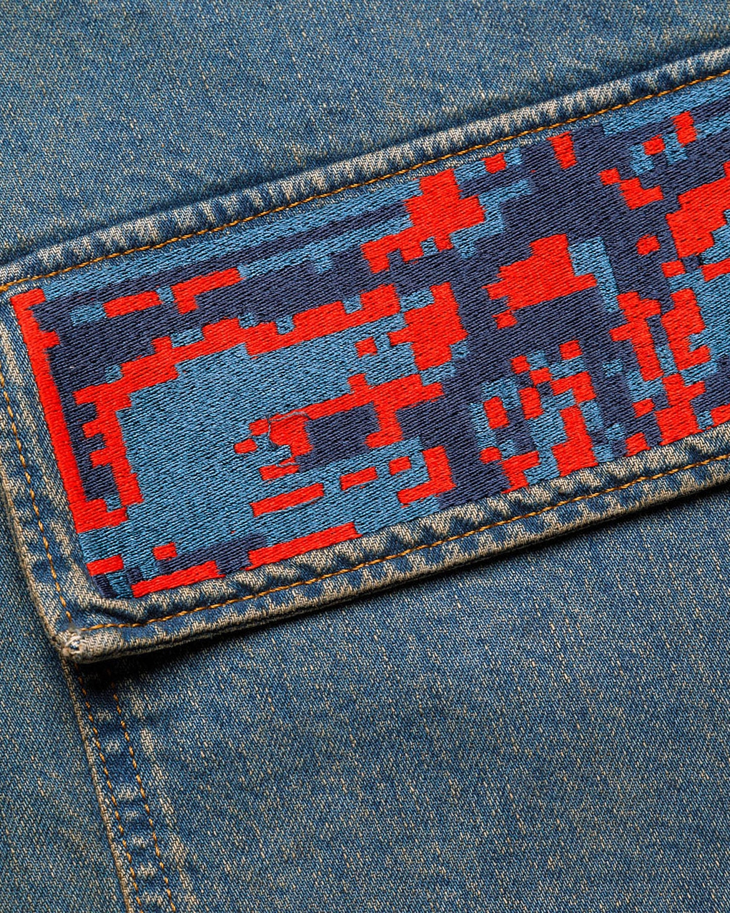 Extreme macro close-up of the pixelated embroidery on the "Coral Biome" denim jacket pocket, showcasing a complex digital glitch pattern in vibrant red and various shades of blue thread.