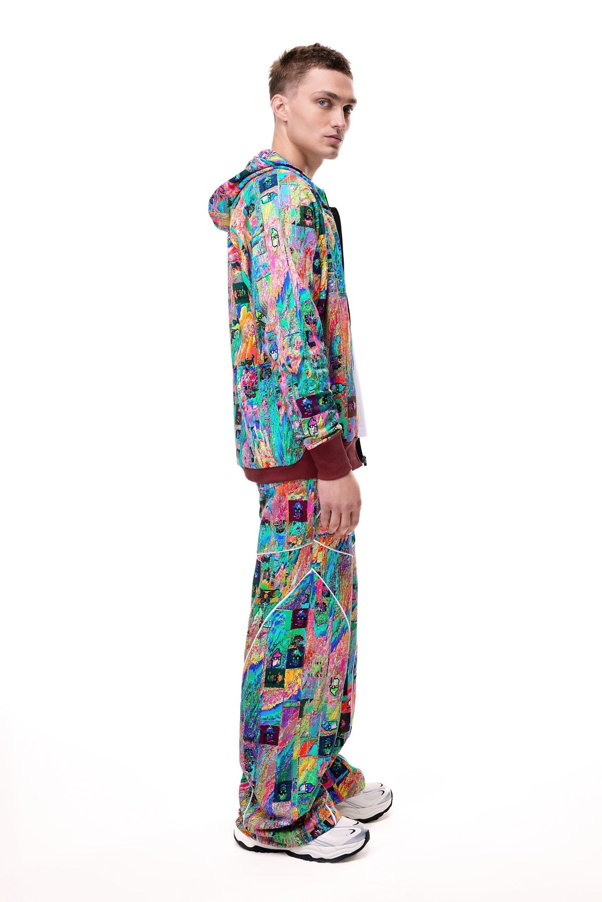 Side profile of a male model wearing the GLIX "Mudderrrfall" full-zipped hoodie and matching trousers, showcasing the fluid multicolor digital glitch print and burgundy ribbed cuffs, styled with white sneakers.