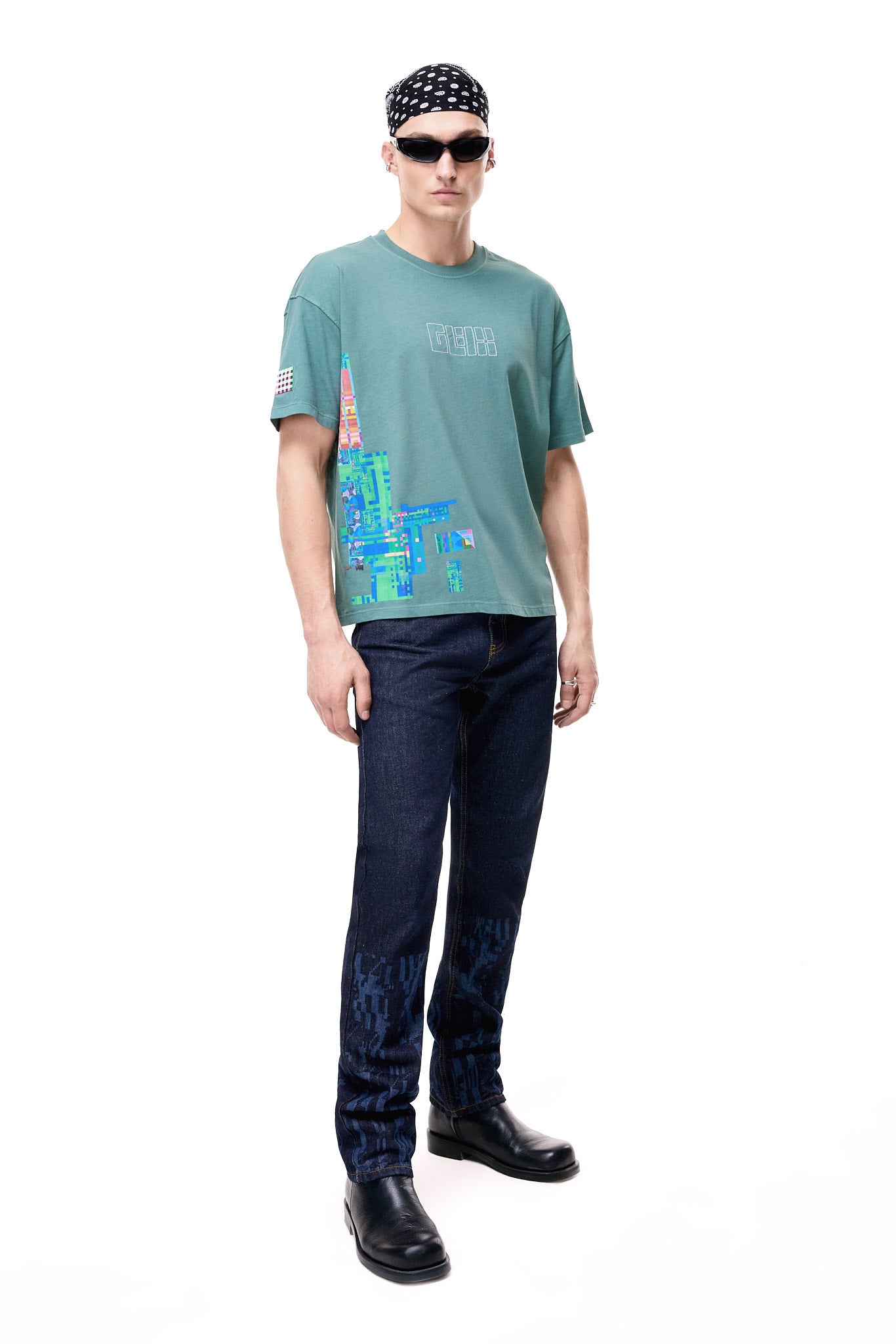 Front view of a male model wearing the green GLIX oversized t-shirt featuring a colorful pixelated glitch graphic on the right side and a white outlined GLIX logo on the chest, styled with dark blue jeans, black boots, and a black bandana.