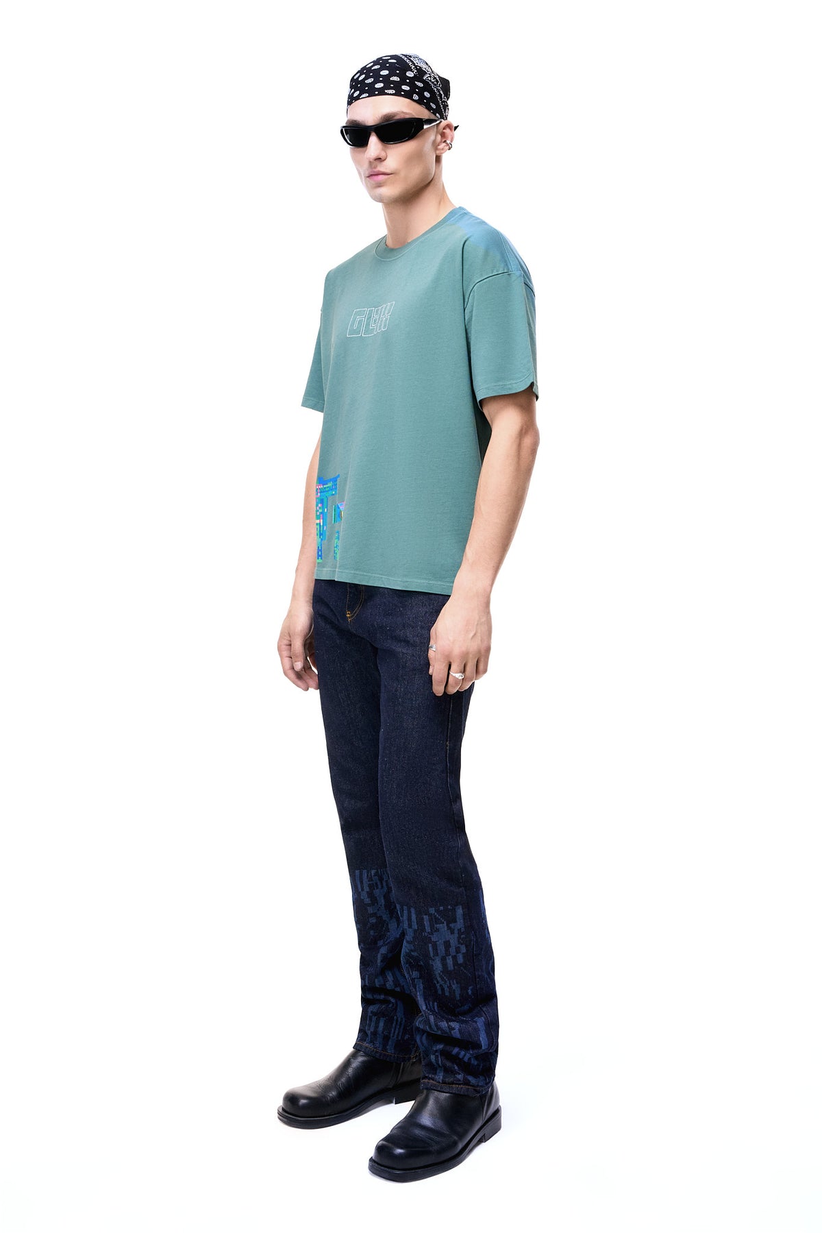 Side view of a male model wearing the green GLIX oversized t-shirt, showing the loose fit and the colorful pixelated glitch graphic wrapping from the front hem, styled with dark denim and black boots.