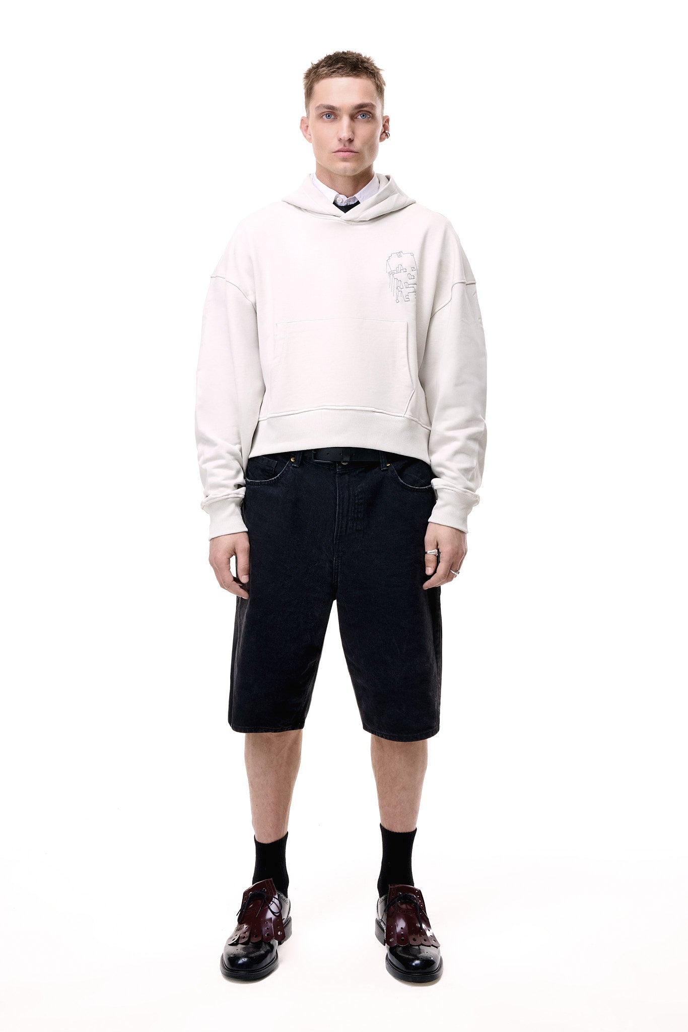 Full-length front view of a male model wearing the light grey GLIX "Grey Matter" oversized hoodie featuring a small grey digital glitch embroidery on the left chest, styled with a white collared shirt, black tie, black denim shorts, and burgundy dress shoes.