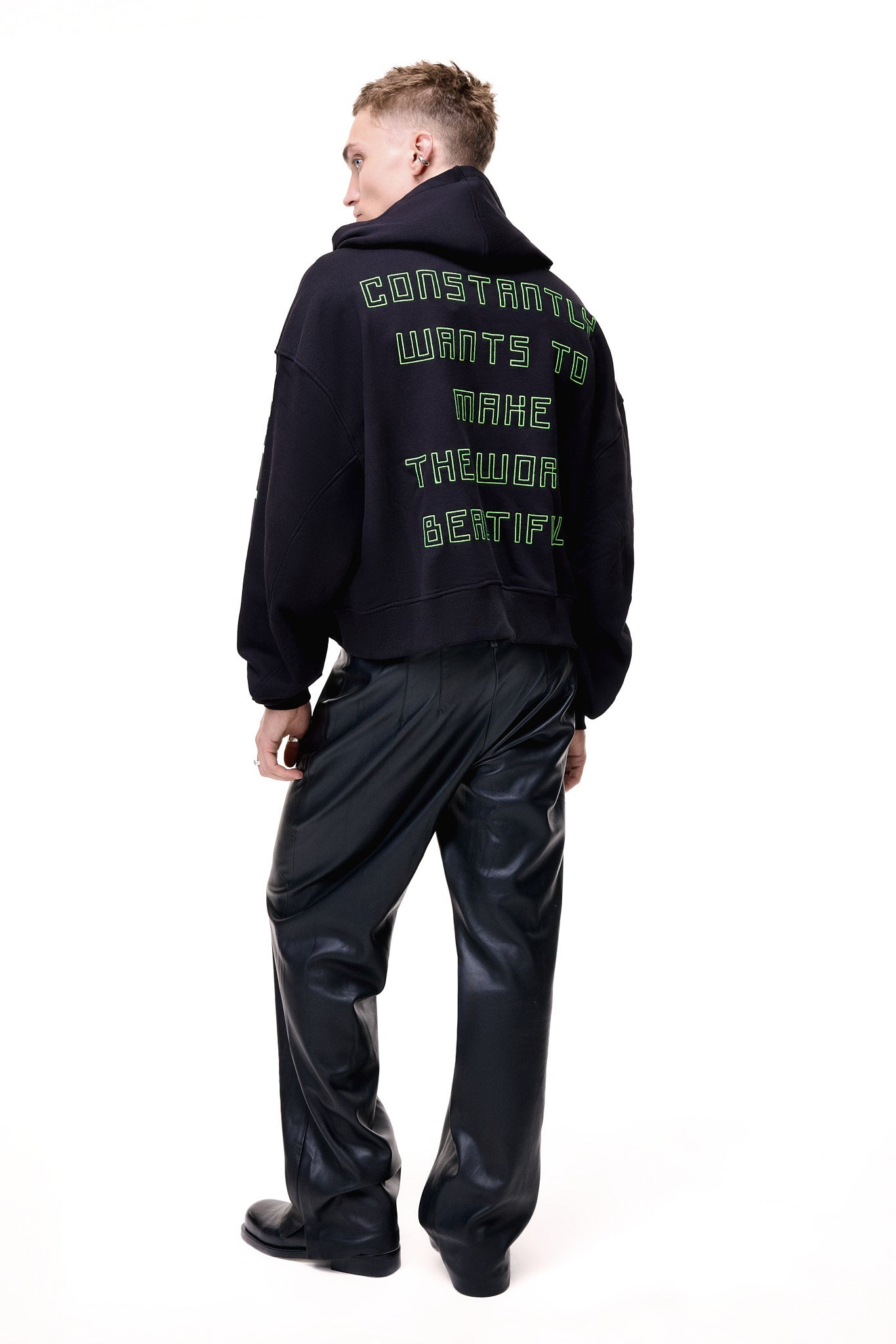 Rear view of a male model wearing the black GLIX "Make It Beautiful" oversized hoodie, featuring neon green puff print text on the back that reads "CONSTANTLY WANTS TO MAKE THE WORLD BEAUTIFUL," styled with black leather trousers.