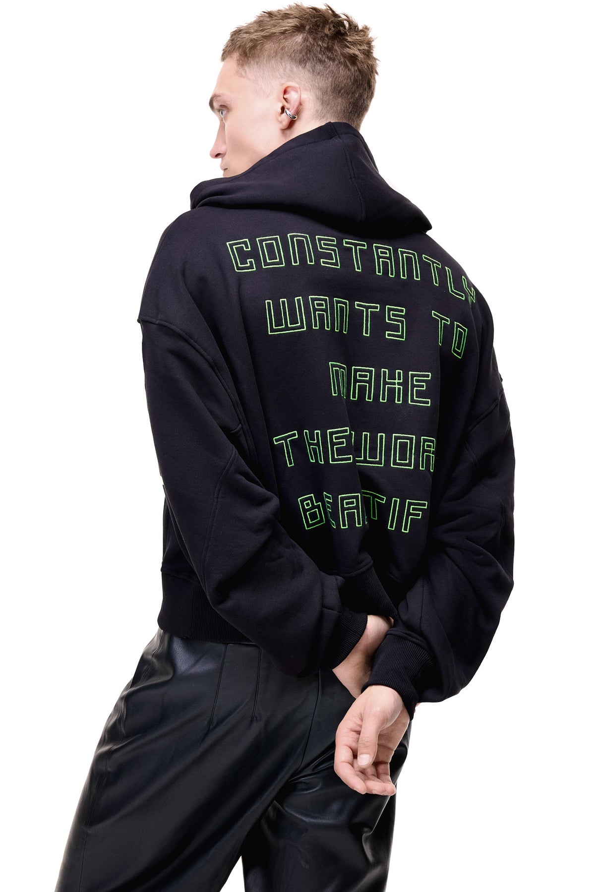 A mid-profile back view of a male model wearing the black GLIX "Make It Beautiful" oversized hoodie, highlighting the neon green puff print typography on the back and the slouchy fit, styled with black leather trousers.