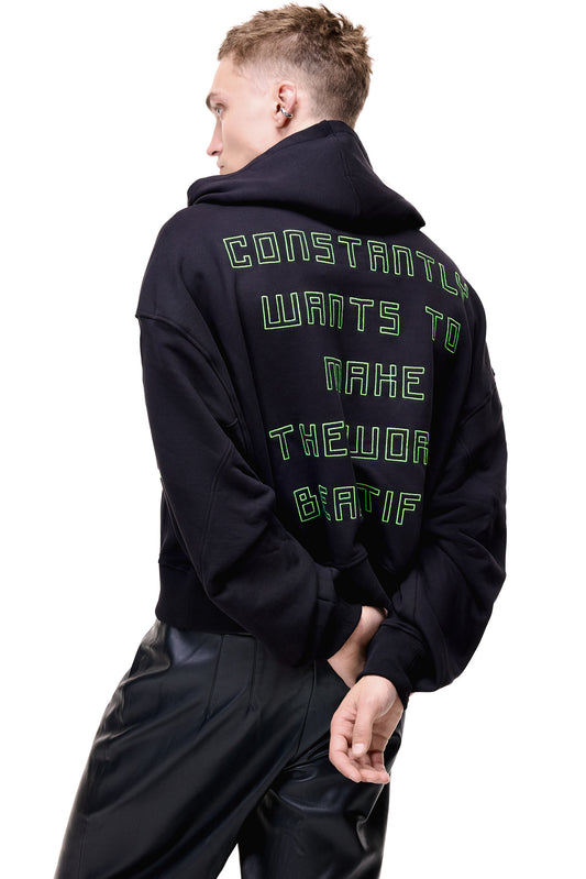 A mid-profile back view of a male model wearing the black GLIX "Make It Beautiful" oversized hoodie, highlighting the neon green puff print typography on the back and the slouchy fit, styled with black leather trousers.