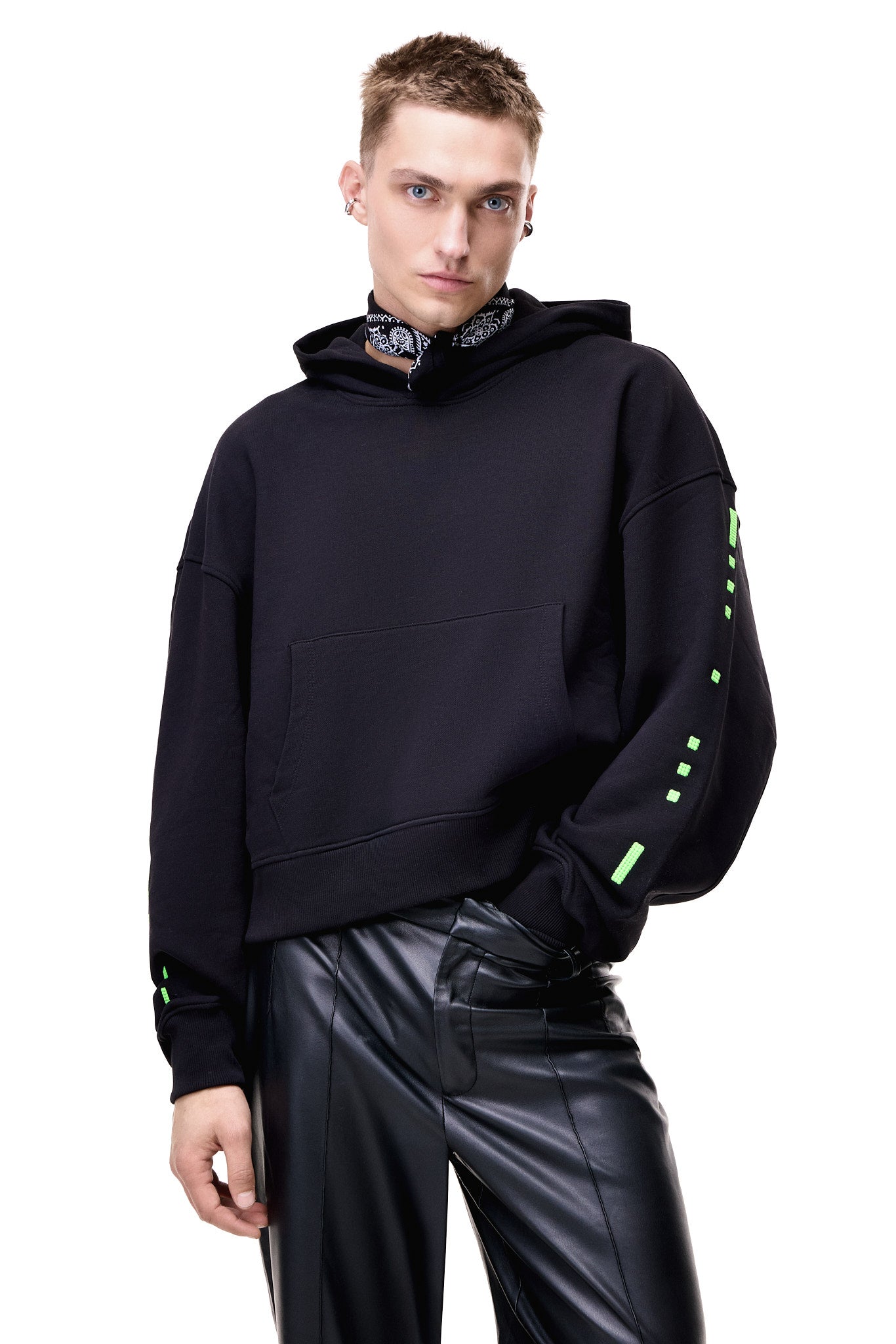 Front view of a male model wearing the black GLIX "Make It Beautiful" oversized hoodie, featuring neon green digital dashed lines on the sleeves and a black and white paisley bandana neck gaiter, styled with black leather trousers.