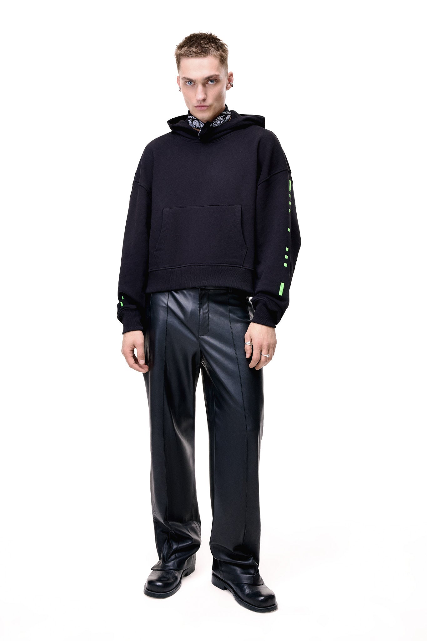 Full-length front view of a male model wearing the black GLIX "Make It Beautiful" oversized hoodie, featuring neon green digital dashed lines on the sleeves, styled with a black and white paisley bandana, black leather trousers, and black leather boots.