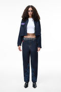 Front lifestyle view of a female model wearing the GLIX "Cumulus" feminine fit jeans in dark wash denim, styled with the matching "Totem" denim crop bomber jacket, a white cropped top, and a silver chain belt.