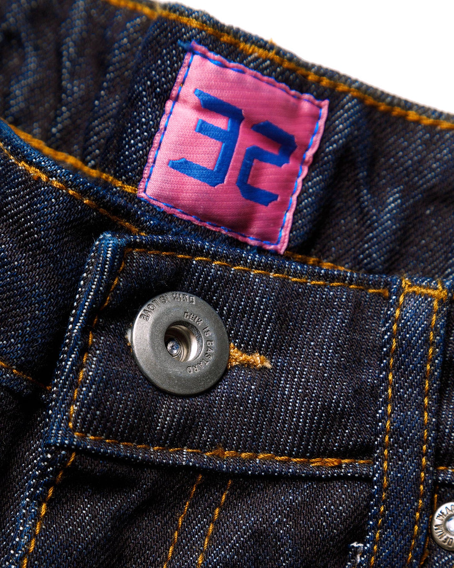 Macro close-up of the front closure on the GLIX "Cumulus" feminine fit jeans, featuring a silver branded button embossed with "GLIX IS BASTARD / GLIX IS LOVE"