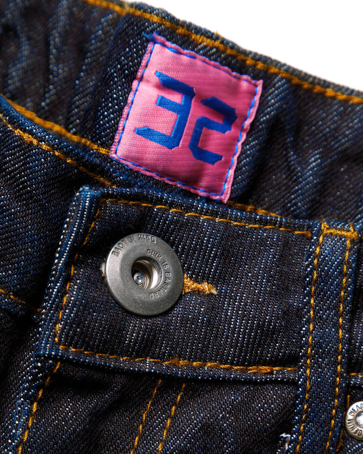 Macro close-up of the front closure on the GLIX "Cumulus" feminine fit jeans, featuring a silver branded button embossed with "GLIX IS BASTARD / GLIX IS LOVE"