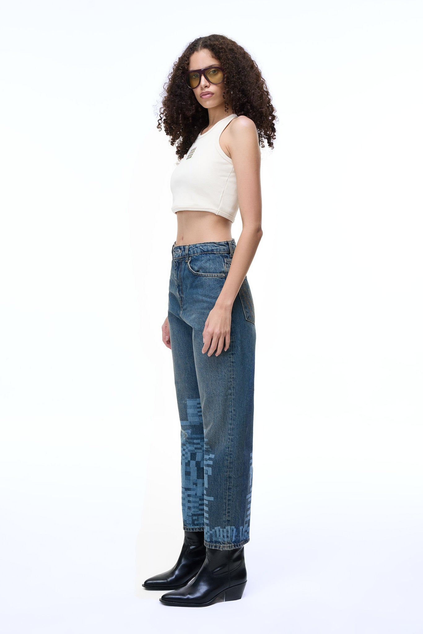 Side profile lifestyle view of a female model wearing the GLIX "Jetpack" feminine fit jeans in mid-wash denim, featuring light blue pixelated glitch graphics on the lower leg, styled with a white ribbed crop top and black leather boots.