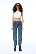 Front lifestyle view of a female model wearing the GLIX "Jetpack" feminine fit jeans in mid-wash denim, featuring light blue pixelated graphics on the lower legs and a red embroidered logo on the coin pocket, styled with a white ribbed crop top and black leather boots.