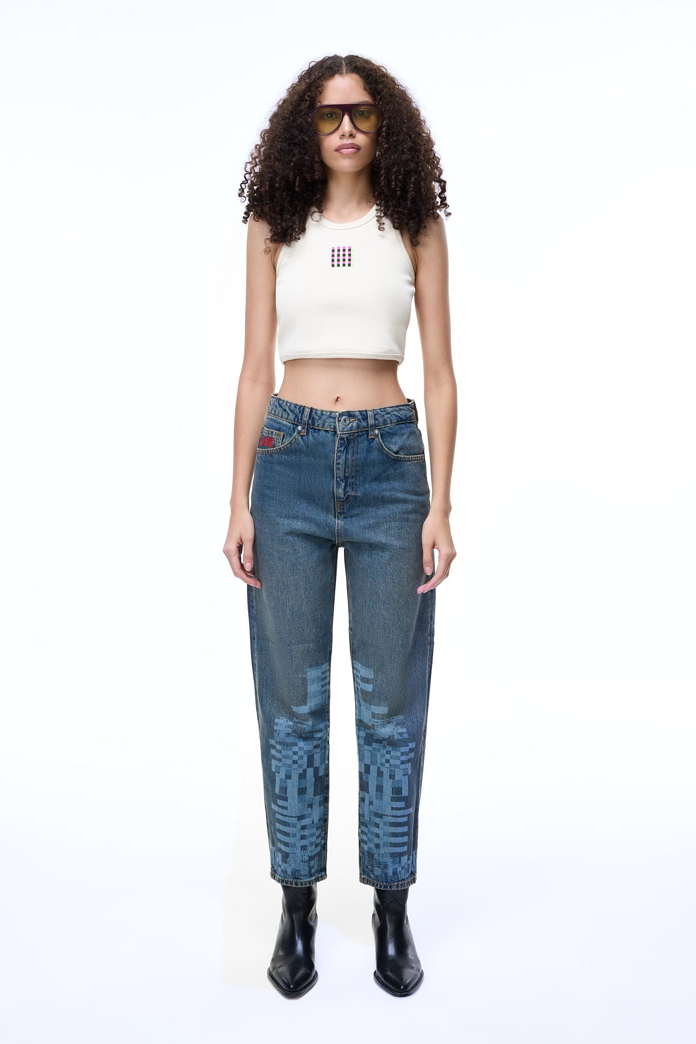 Front lifestyle view of a female model wearing the GLIX "Jetpack" feminine fit jeans in mid-wash denim, featuring light blue pixelated graphics on the lower legs and a red embroidered logo on the coin pocket, styled with a white ribbed crop top and black leather boots.