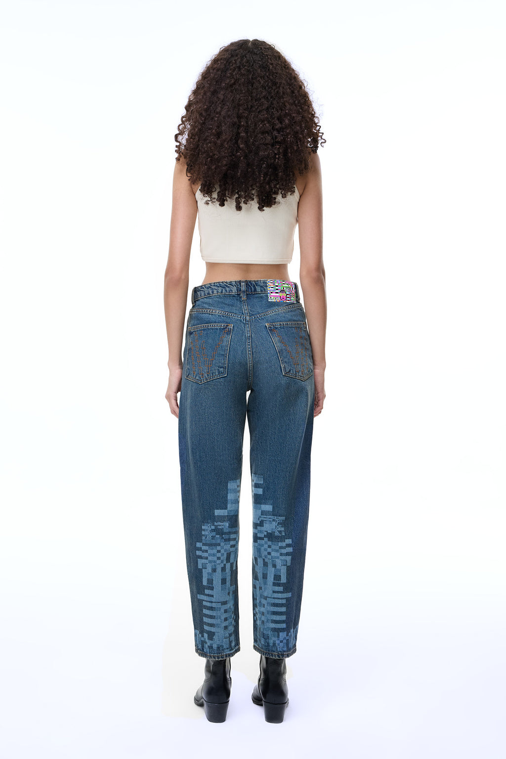 Macro close-up of the back pocket on the GLIX "Jetpack" feminine fit jeans, featuring a distinct black pixelated "V" embroidery design on mid-wash denim with contrast orange stitching.