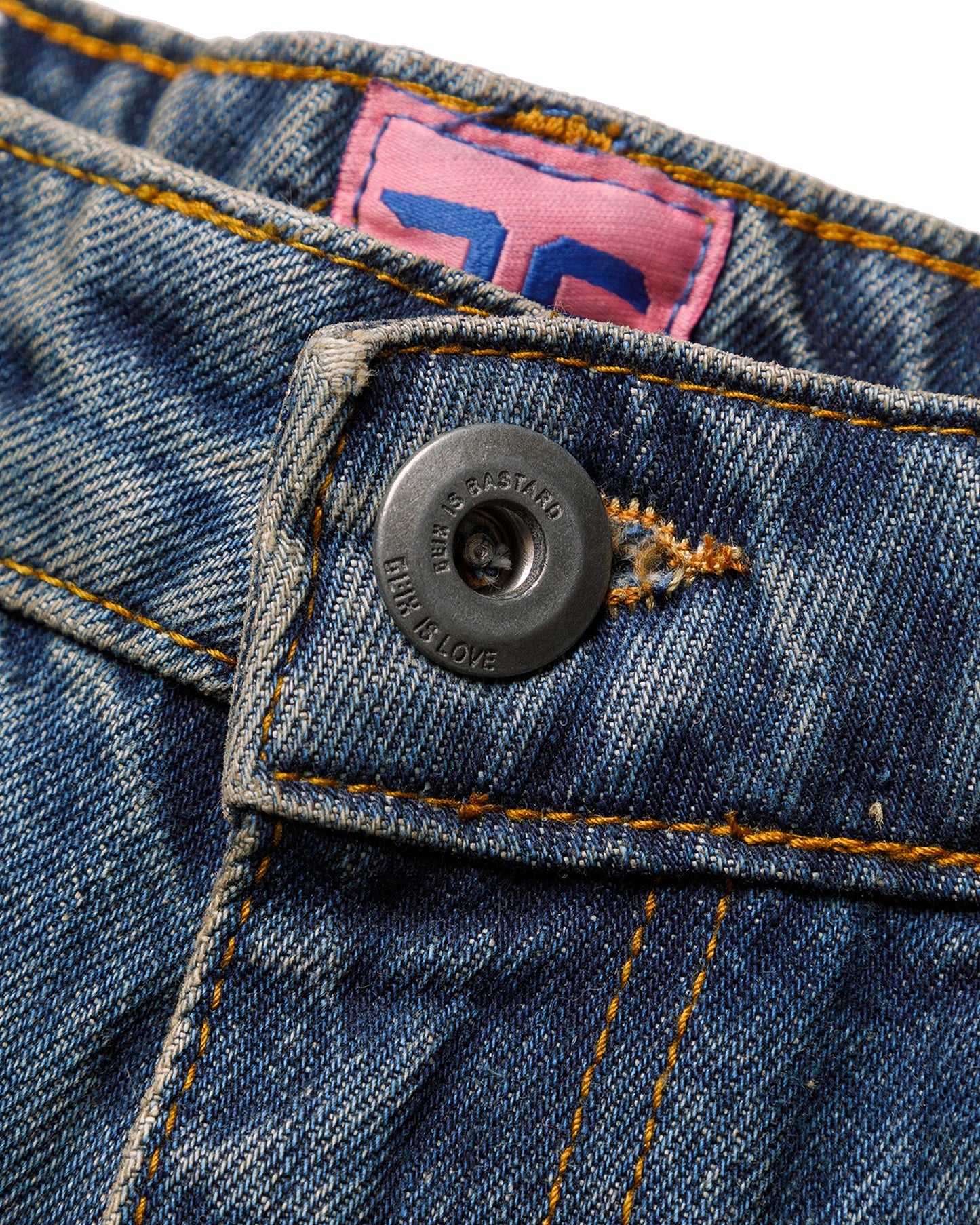 Macro close-up of the front closure on the GLIX "Jetpack" feminine fit jeans, featuring a silver branded button embossed with "GLIX IS BASTARD / GLIX IS LOVE" and a pink internal size tag with a blue graphic on mid-wash denim.