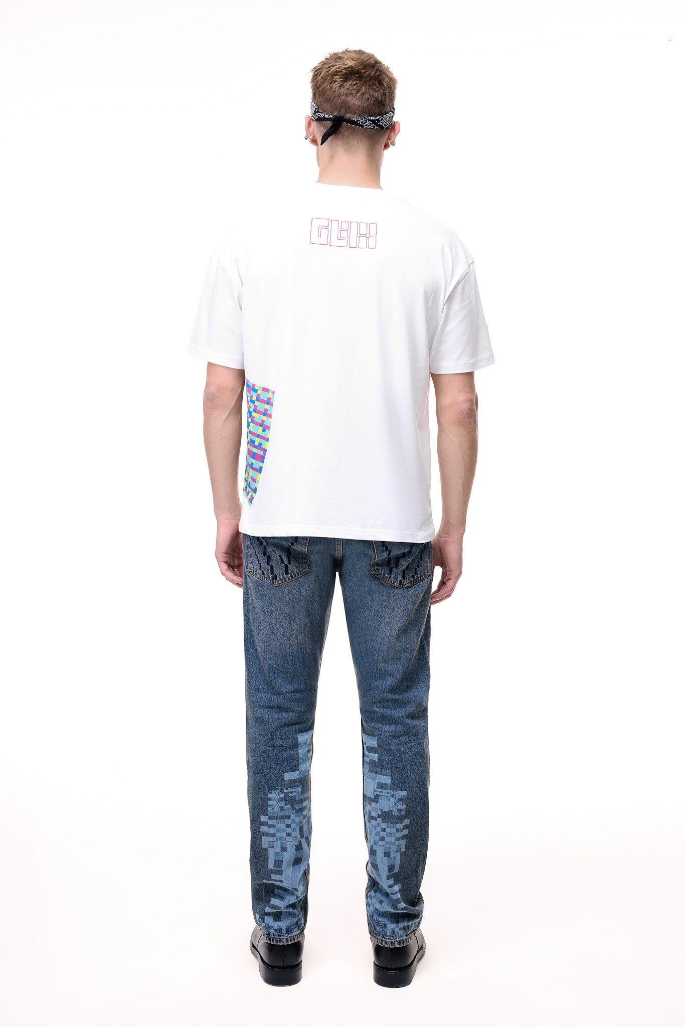 Rear lifestyle view of a male model wearing the GLIX "Jetpack" mid-wash denim jeans, showcasing light blue pixelated digital glitch graphics on the back of the lower legs and classic five-pocket construction with contrast orange stitching.