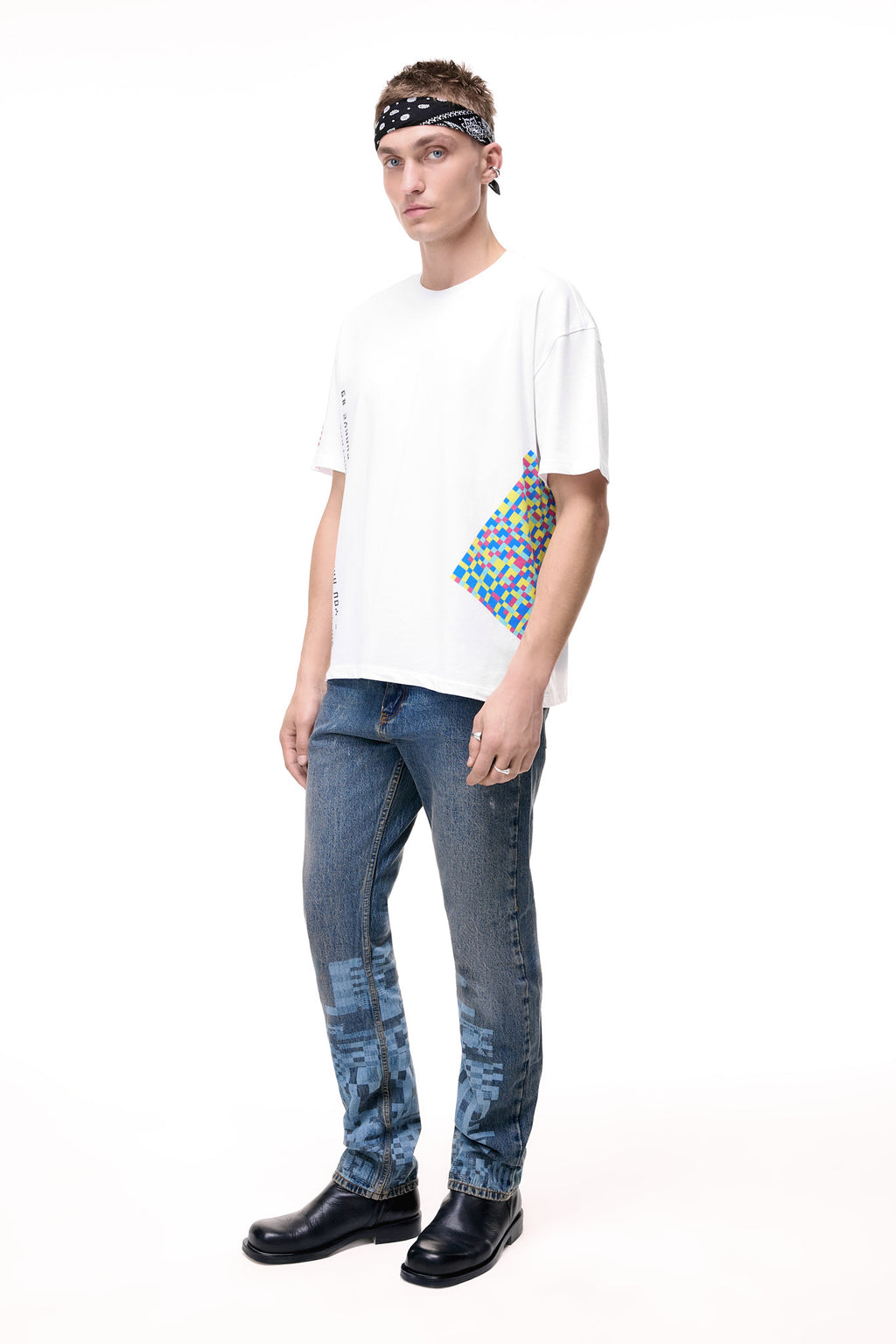 Front lifestyle view of a male model wearing the GLIX "Jetpack" mid-wash denim jeans in a masculine fit, showcasing light blue pixelated glitch graphics on the lower legs, styled with a white graphic t-shirt and a black and white paisley headband.