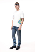 Front lifestyle view of a male model wearing the GLIX "Jetpack" mid-wash denim jeans in a masculine fit, showcasing light blue pixelated glitch graphics on the lower legs, styled with a white graphic t-shirt and a black and white paisley headband.