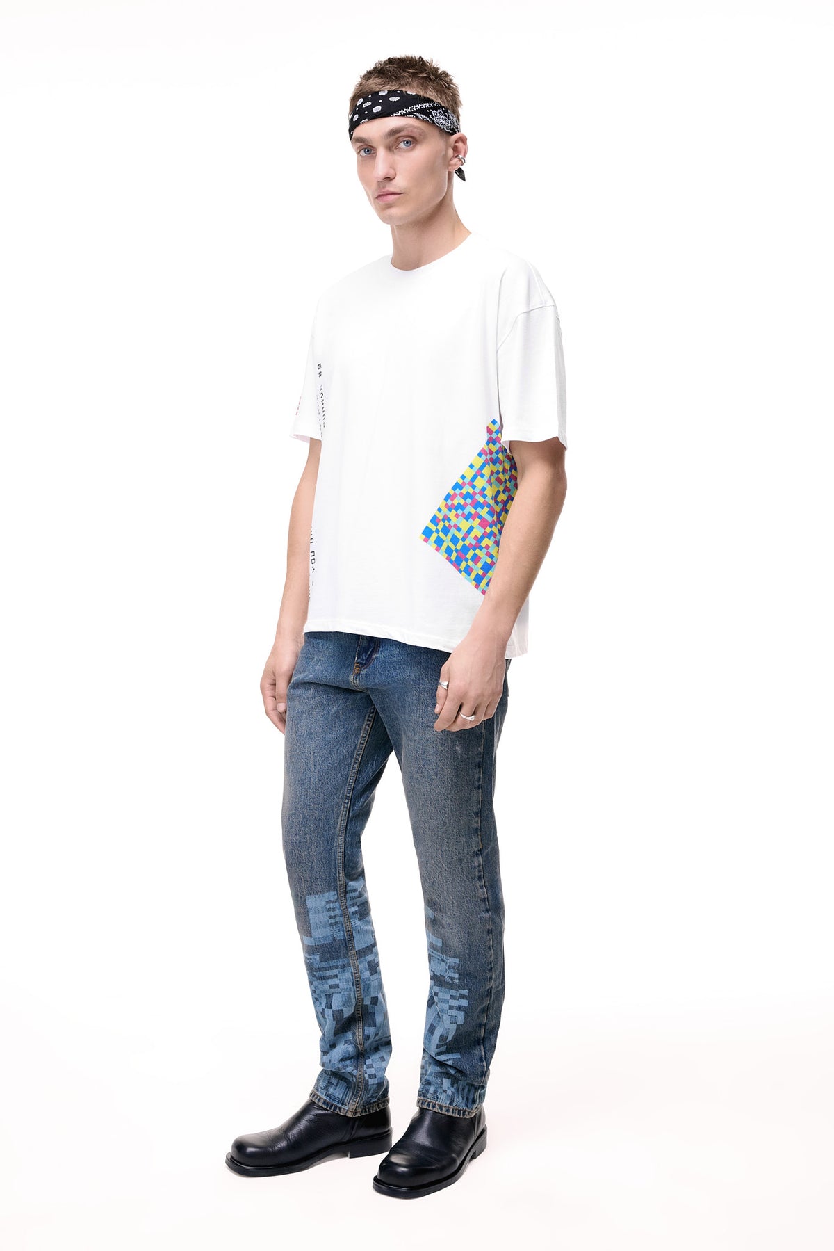 Front lifestyle view of a male model wearing the GLIX "Jetpack" mid-wash denim jeans in a masculine fit, showcasing light blue pixelated glitch graphics on the lower legs, styled with a white graphic t-shirt and a black and white paisley headband.