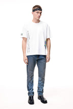 Front lifestyle view of a male model wearing the GLIX "Jetpack" mid-wash denim jeans in a masculine fit, featuring light blue pixelated digital glitch graphics on the lower legs, styled with a white graphic t-shirt and a black paisley headband.