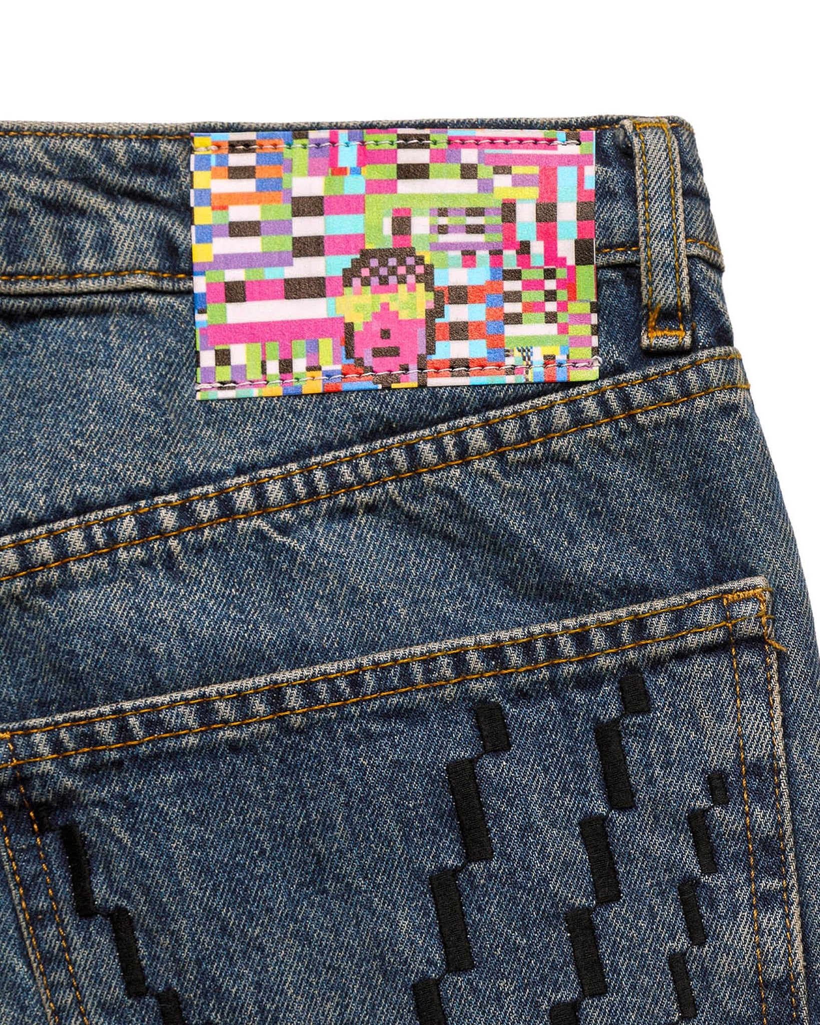 Macro close-up of the GLIX "Jetpack" jeans' back waist patch, featuring a vibrant, multi-colored digital glitch pattern with a pixelated character head in pink and yellow on mid-wash denim.