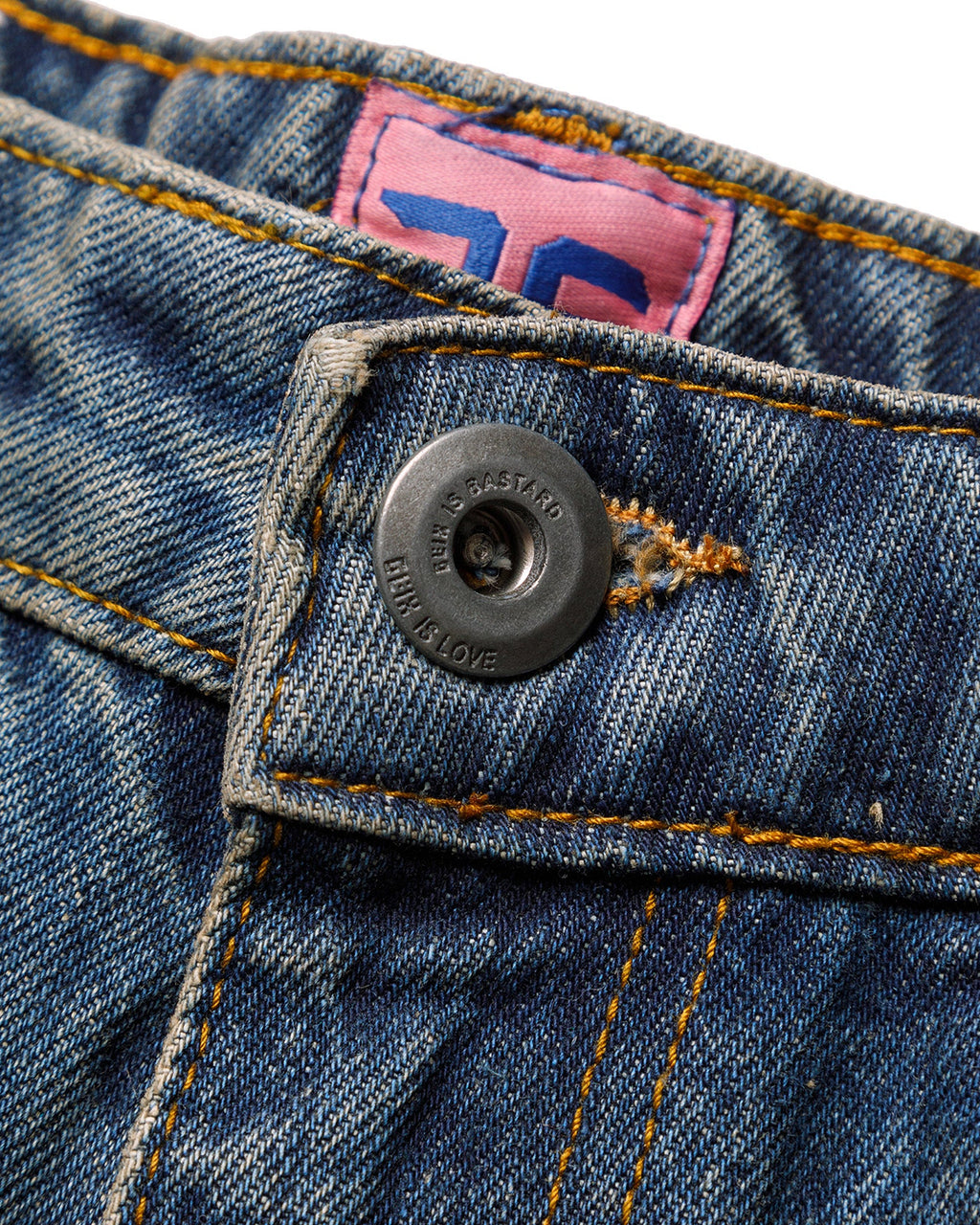 Macro close-up of the front closure on the GLIX "Jetpack" masculine fit jeans, featuring a silver branded button embossed with "GLIX IS BASTARD / GLIX IS LOVE"