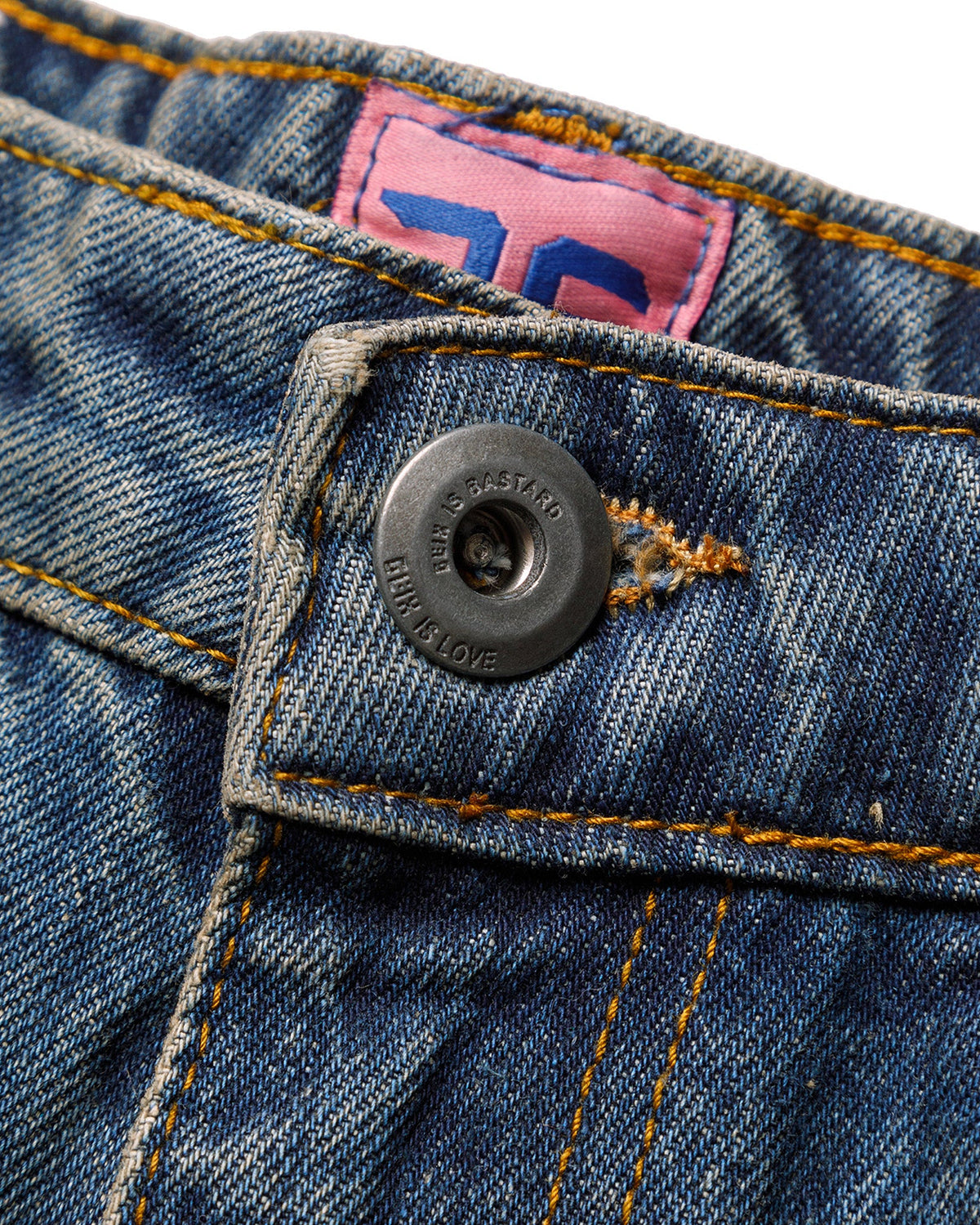 Macro close-up of the front closure on the GLIX "Jetpack" masculine fit jeans, featuring a silver branded button embossed with "GLIX IS BASTARD / GLIX IS LOVE"