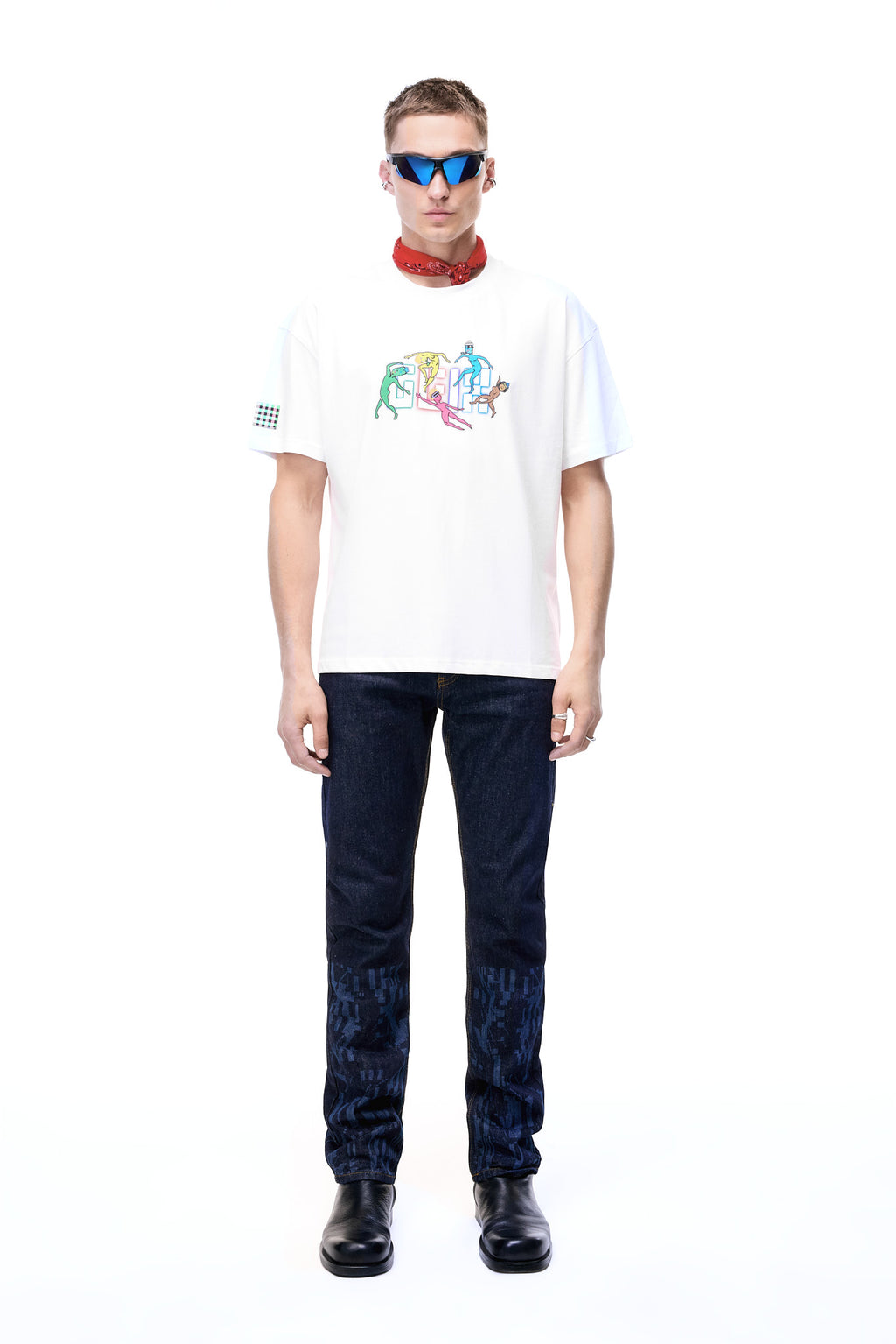 Front lifestyle view of a male model wearing the GLIX "The Maze" dark wash denim jeans in a masculine fit, featuring tonal blue pixelated maze graphics on the lower legs, styled with a white graphic t-shirt, red neck bandana, and blue-tinted sunglasses.