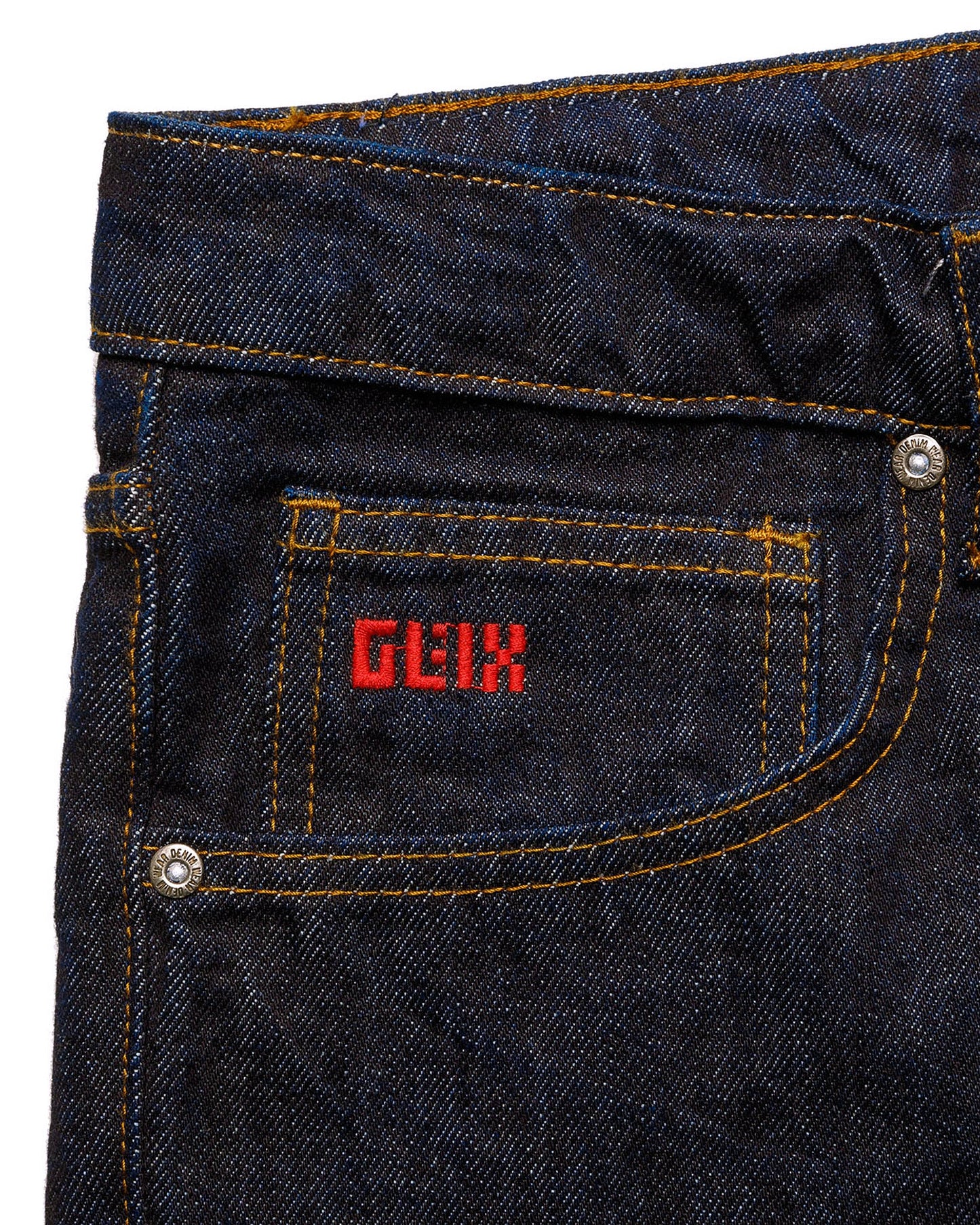 Macro close-up of the front pocket on the GLIX "The Maze" masculine fit jeans, featuring a small red embroidered GLIX logo on the coin pocket and silver branded rivets on dark wash denim.