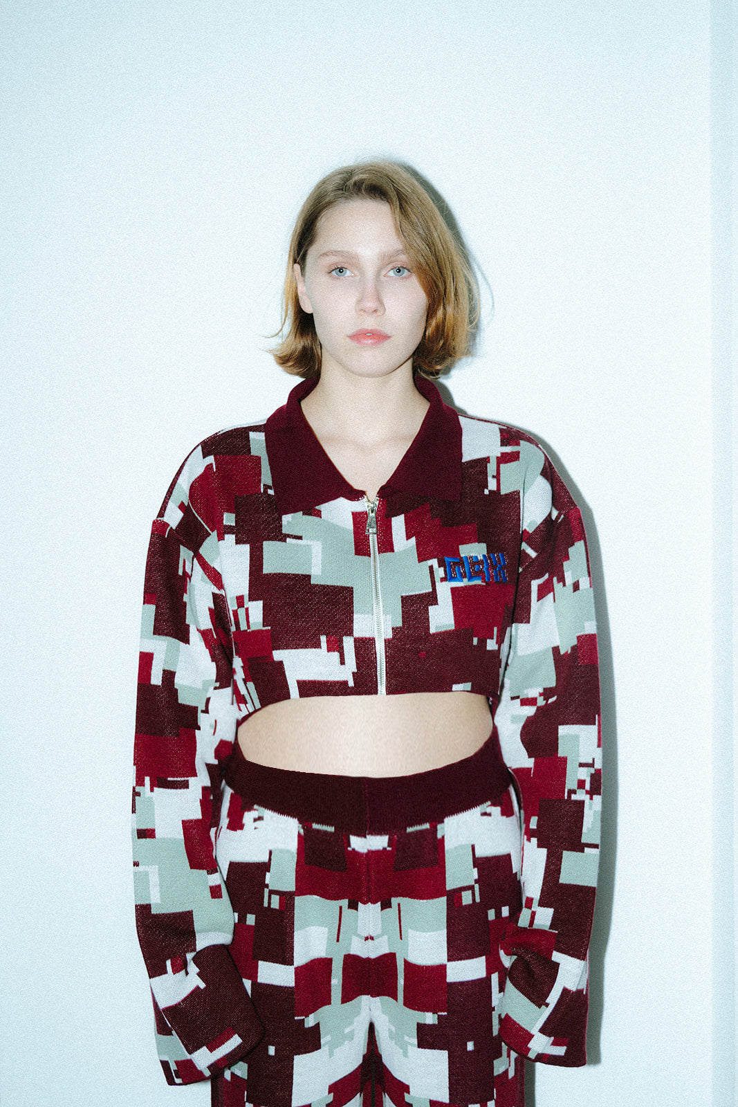 Front view of a female model wearing the GLIX "Cherry Moo" cropped zip-up jacquard knit jacket in burgundy, sage green, and white pixelated glitch pattern, with blue embroidered GLIX logo on the chest and solid burgundy polo collar, styled with matching Cherry Moo wide-leg knit trousers against a white studio background.