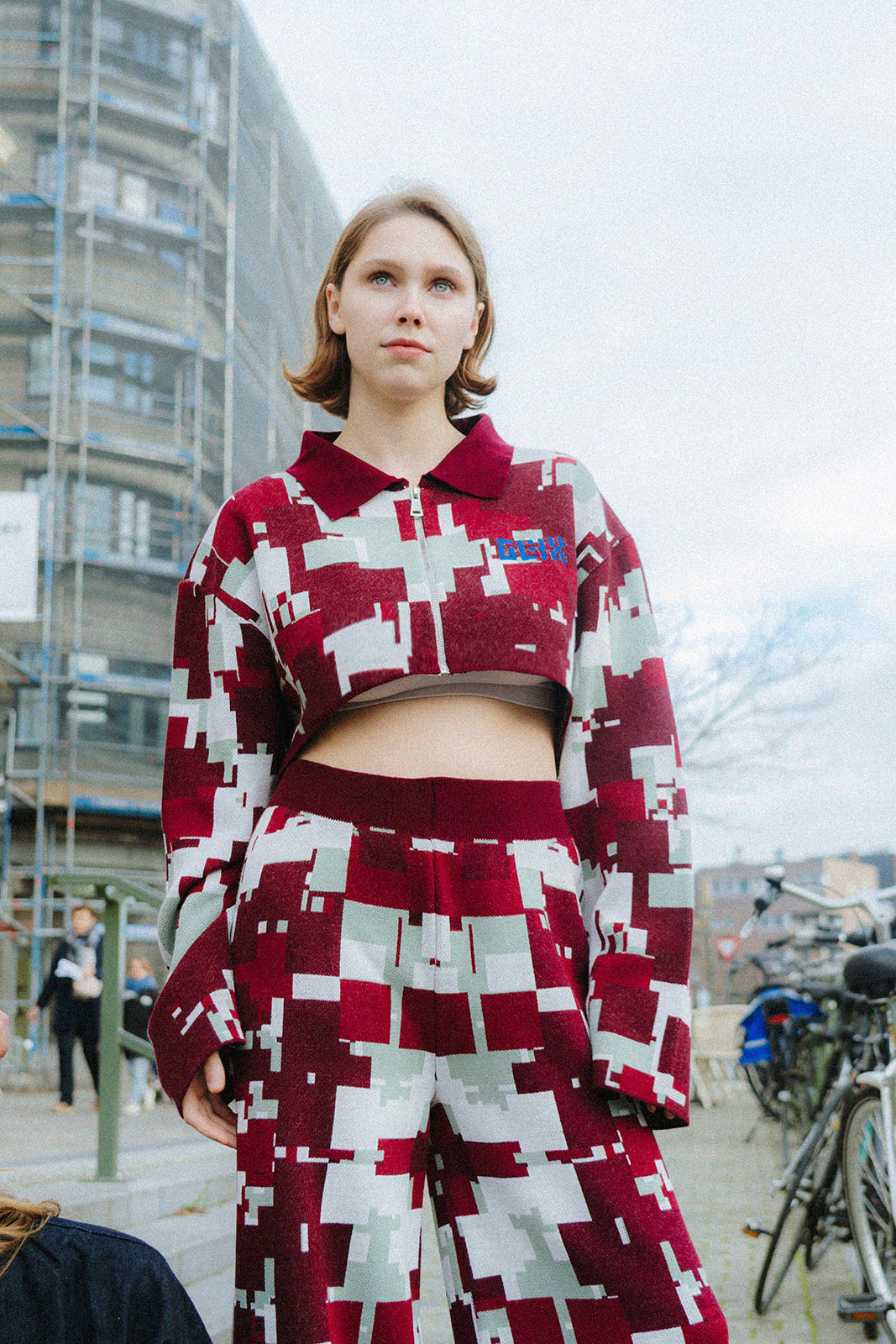 Full-length front view of a female model wearing the GLIX "Cherry Moo" cropped jacquard knit jacket and matching wide-leg trousers in burgundy, sage green, and white pixelated glitch pattern, with blue embroidered GLIX logo, styled with black boots on a city street with bicycles and brutalist architecture in the background.
