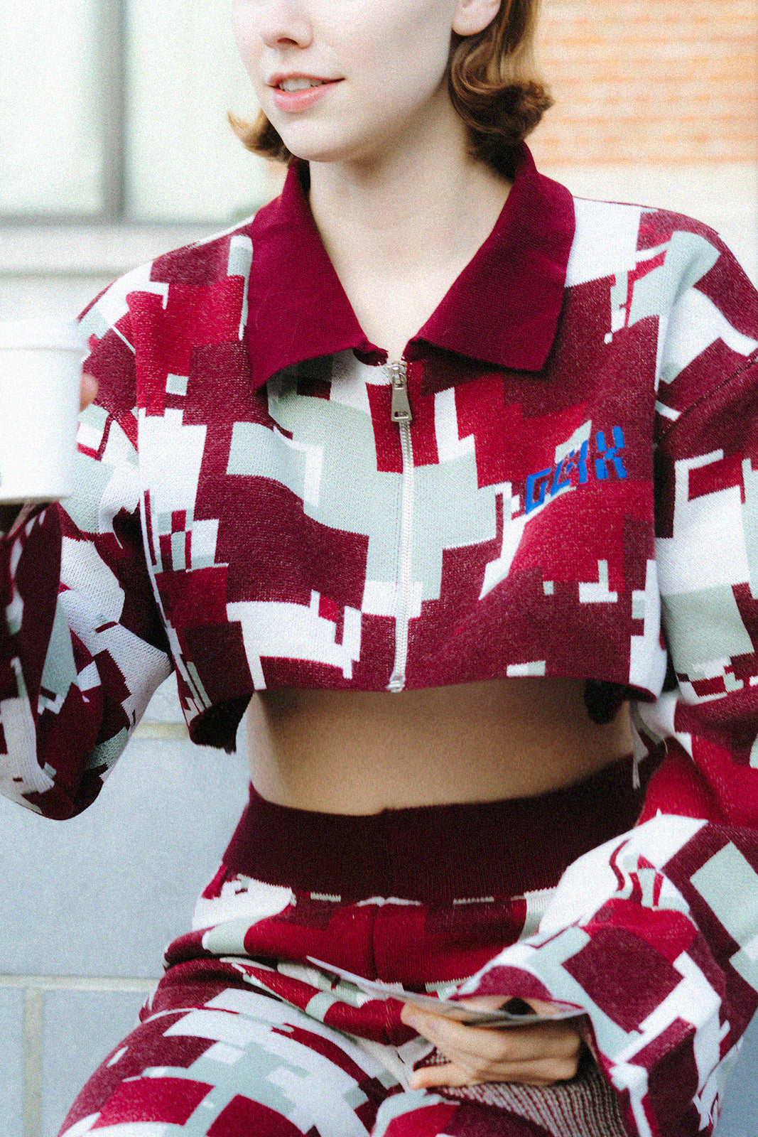 Upper-body close-up of a female model wearing the GLIX "Cherry Moo" cropped zip-up jacquard knit jacket, highlighting the silver centre zip, solid burgundy polo collar, blue embroidered GLIX logo, and the all-over pixelated glitch pattern in burgundy, sage green, and white, with matching wide-leg knit trousers visible below.