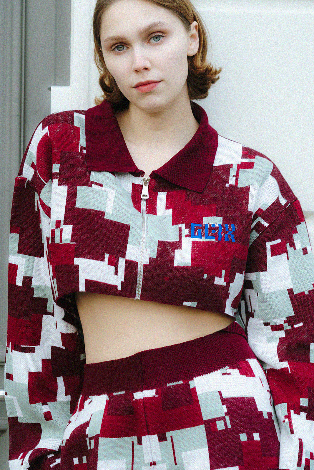 Front three-quarter view of a female model wearing the GLIX "Cherry Moo" cropped zip-up jacquard knit jacket with blue embroidered GLIX logo and solid burgundy polo collar, paired with matching Cherry Moo wide-leg knit trousers in burgundy, sage green, and white pixelated glitch pattern.