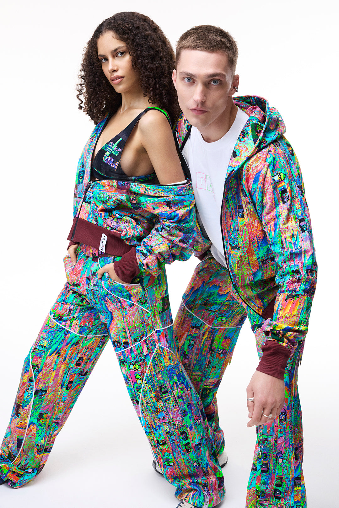 Two models wearing the GLIX MUDDERRRFALL matching set, featuring a colorful generative glitch art pattern on a full zipped hoodie and oversized sweatpants against a white background.