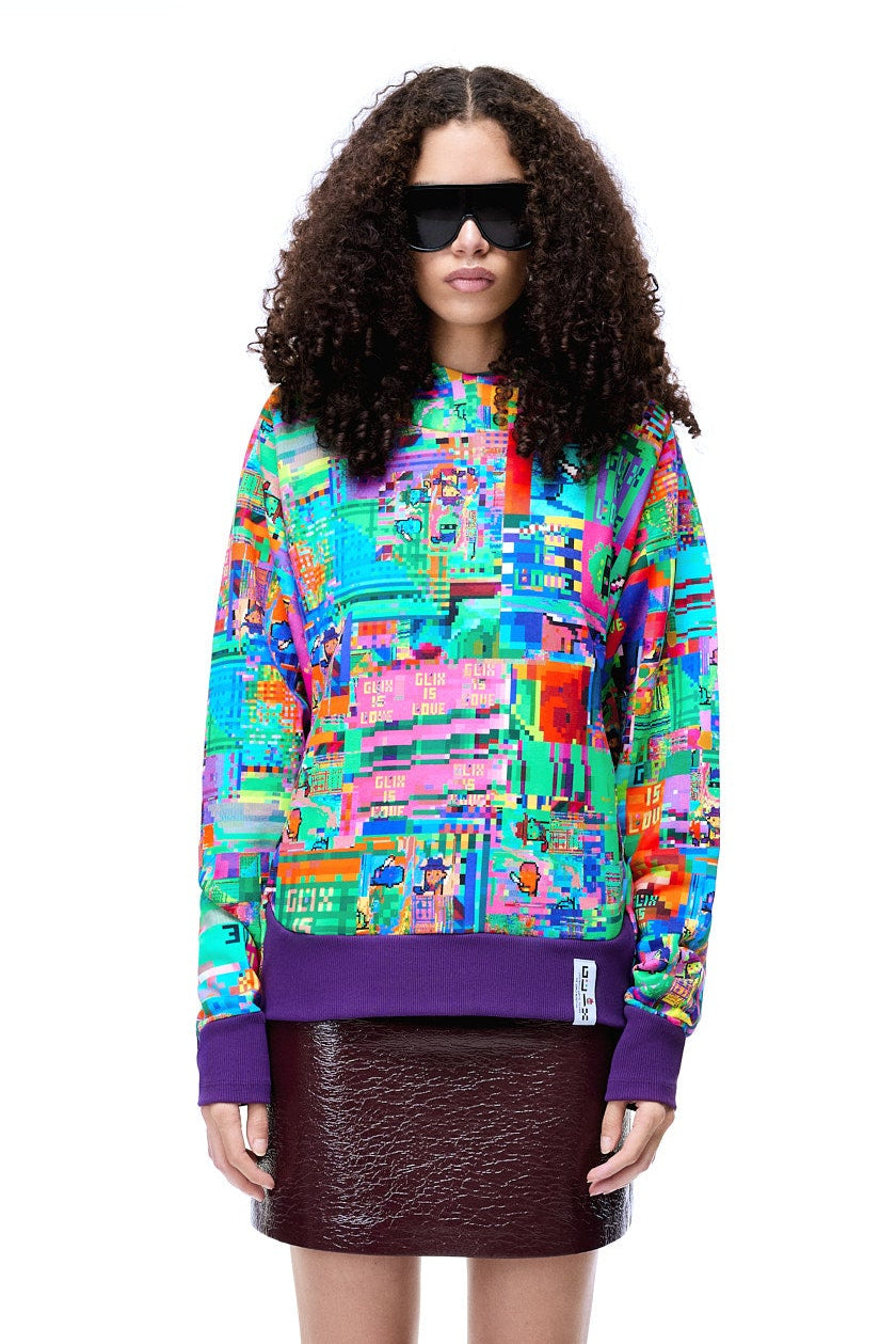 Front view of a female model with curly hair wearing the GLIX "Glix Is Love" oversized hoodie, featuring a vibrant multicolor digital glitch all-over print and purple ribbed hem, styled with large black sunglasses and a dark textured mini skirt.