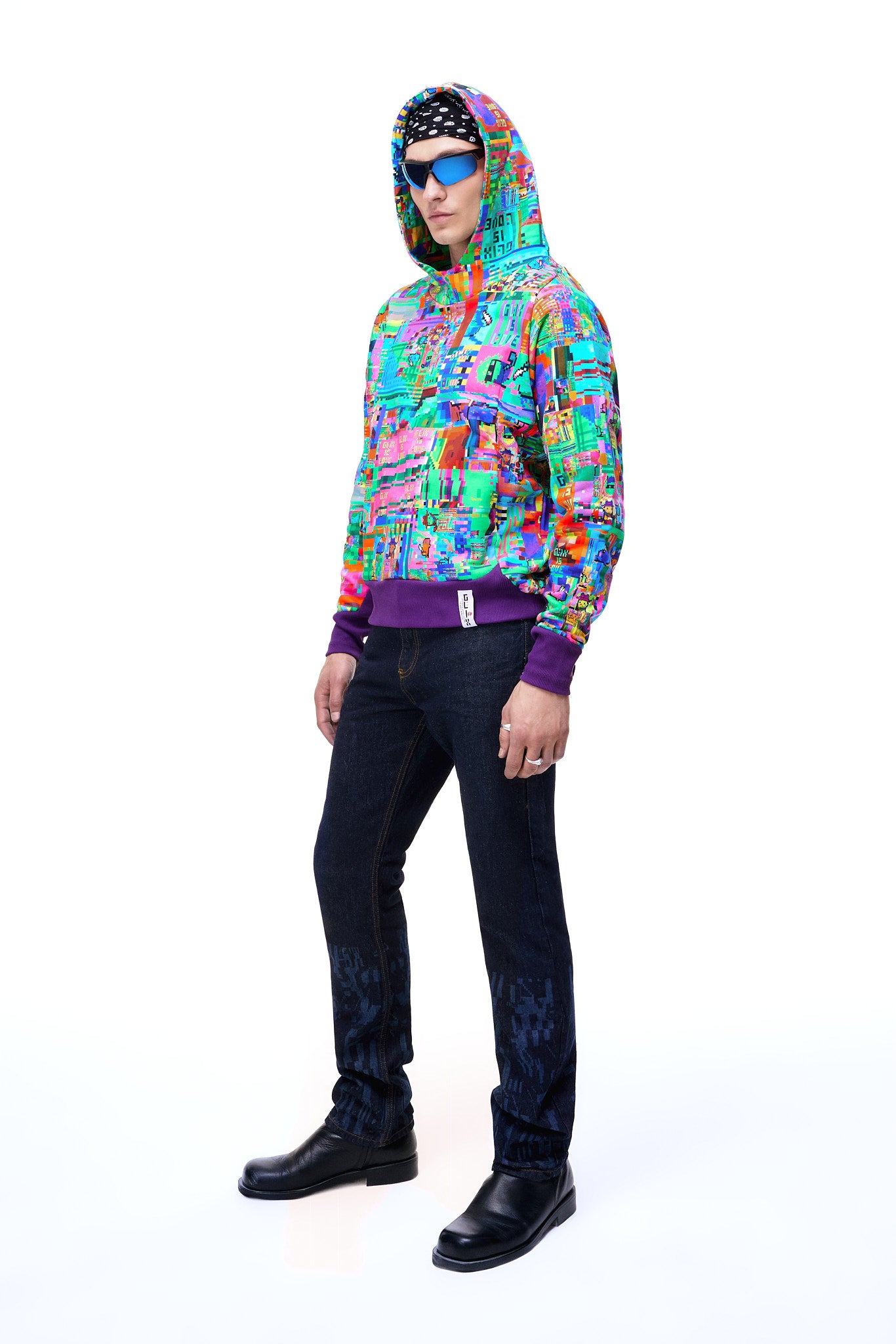 Side profile of a male model wearing the GLIX "Glix Is Love" oversized hoodie with the hood up over a bandana, showcasing the vibrant multicolor digital glitch print on the sleeves and torso, paired with dark blue jeans and black boots.